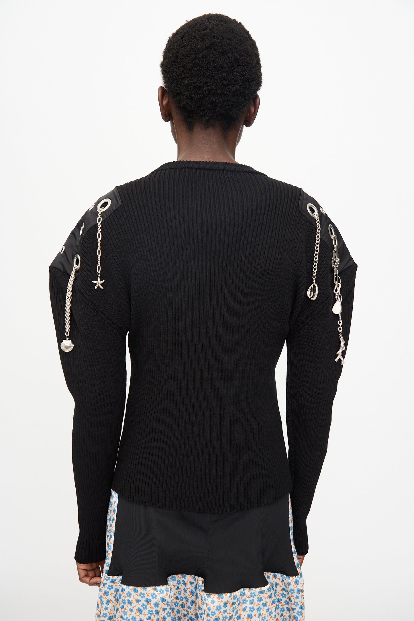 Person facing away, wearing the TOGA ARCHIVES Decoration Rib Knit Pullover in Black featuring metal chains and charms on the shoulders, styled with a patterned skirt.