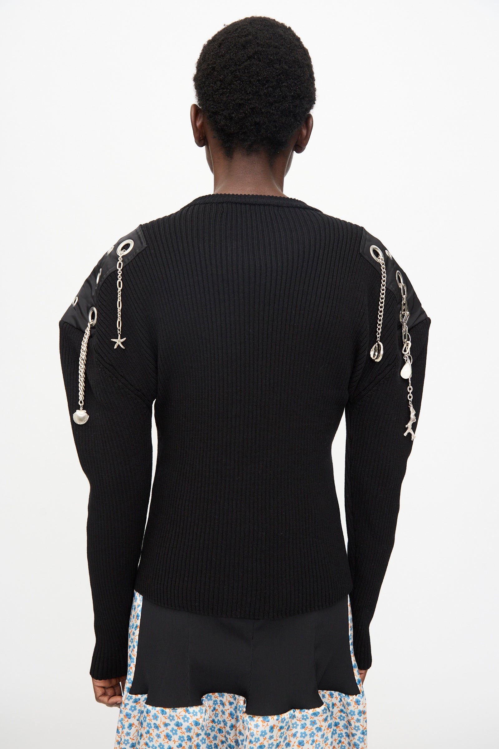 Person facing away, wearing the TOGA ARCHIVES Decoration Rib Knit Pullover in Black featuring metal chains and charms on the shoulders, styled with a patterned skirt.