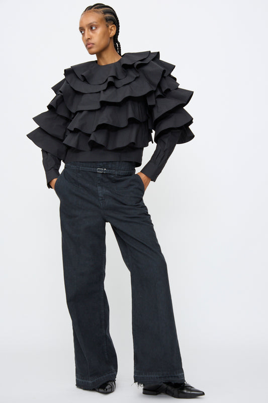 A person wearing a black, long-sleeve, multi-layered ruffled top and TOGA ARCHIVES Denim Pants in Black stands against a plain white background.