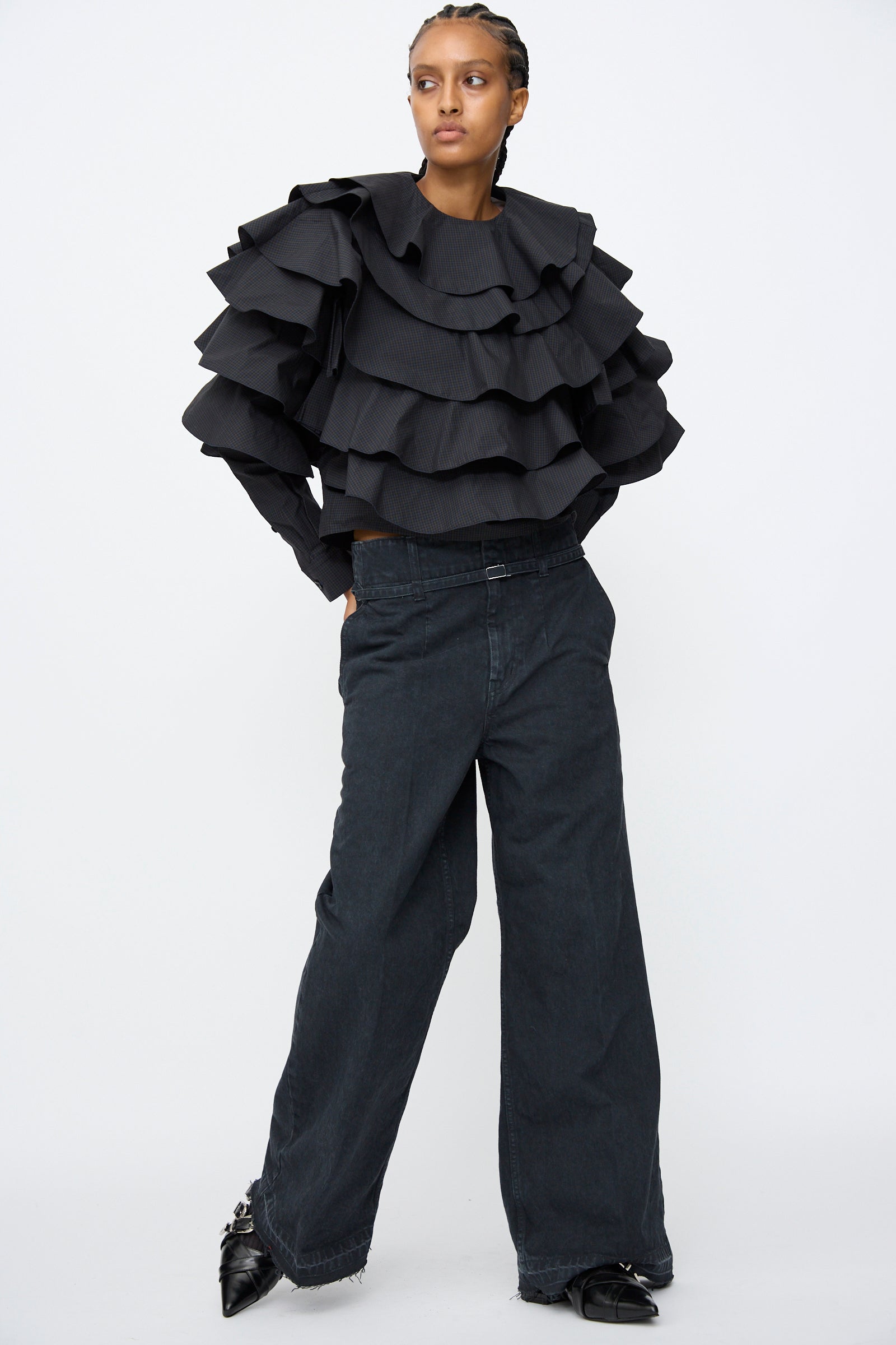 A person stands against a white background wearing TOGA ARCHIVES' Denim Pants in Black, featuring a flared leg and high waist, paired with a black ruffled layered top and one hand in their pocket.