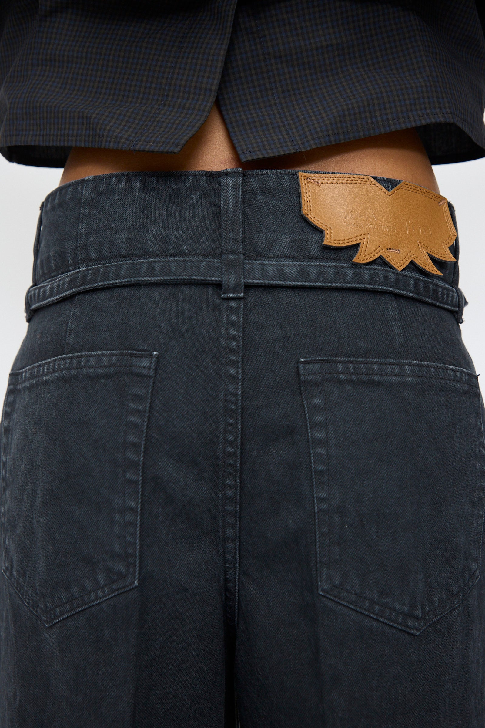 Rear view of a person wearing TOGA ARCHIVES Denim Pants in Black, featuring high waist, flared legs, visible back pockets, and a brown leather patch on the waistband.