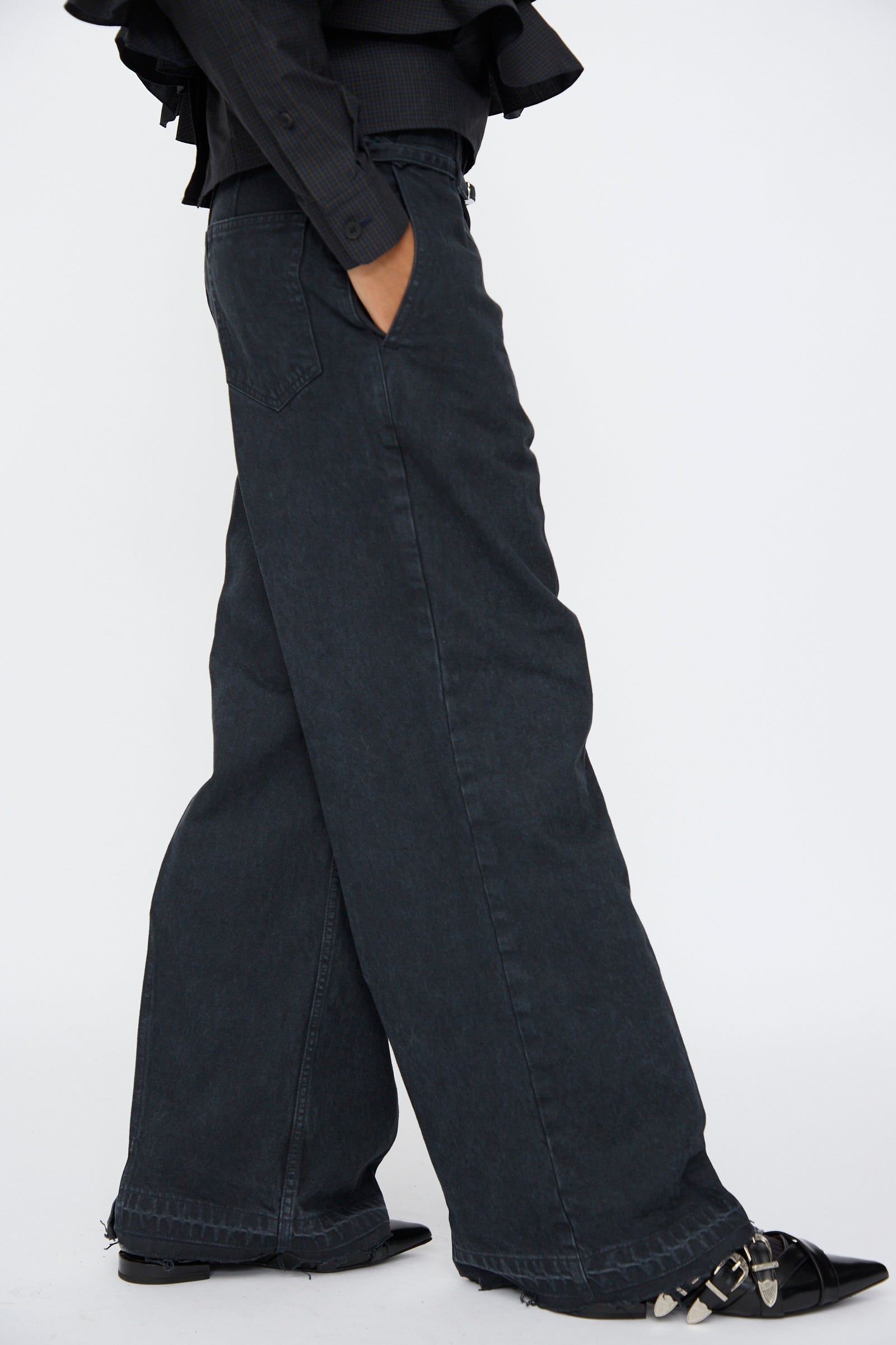 Model wearing TOGA ARCHIVES Denim Pants in Black with a high waist and flared legs, styled with a black long-sleeve shirt and black pointed shoes, photographed from the side with hands in pockets.