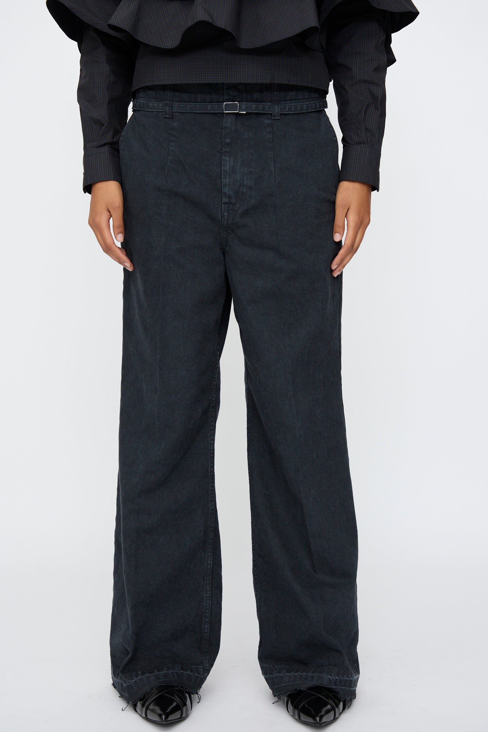 A person wears TOGA ARCHIVES Denim Pants in Black with a flared leg, a belt, black shoes, and a ruffled black long-sleeve top, standing against a plain white background.