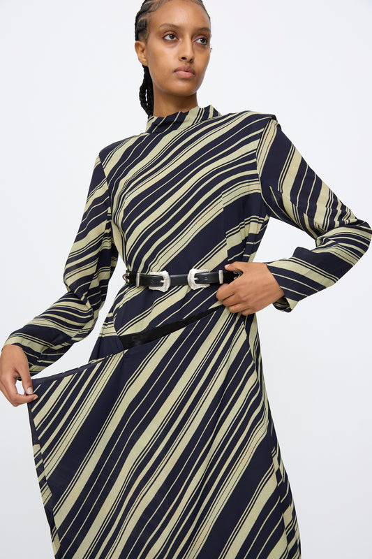 A person stands against a plain white background wearing a long-sleeved black dress with diagonal beige stripes, styled with the TOGA ARCHIVES Double Buckle Belt in Black.