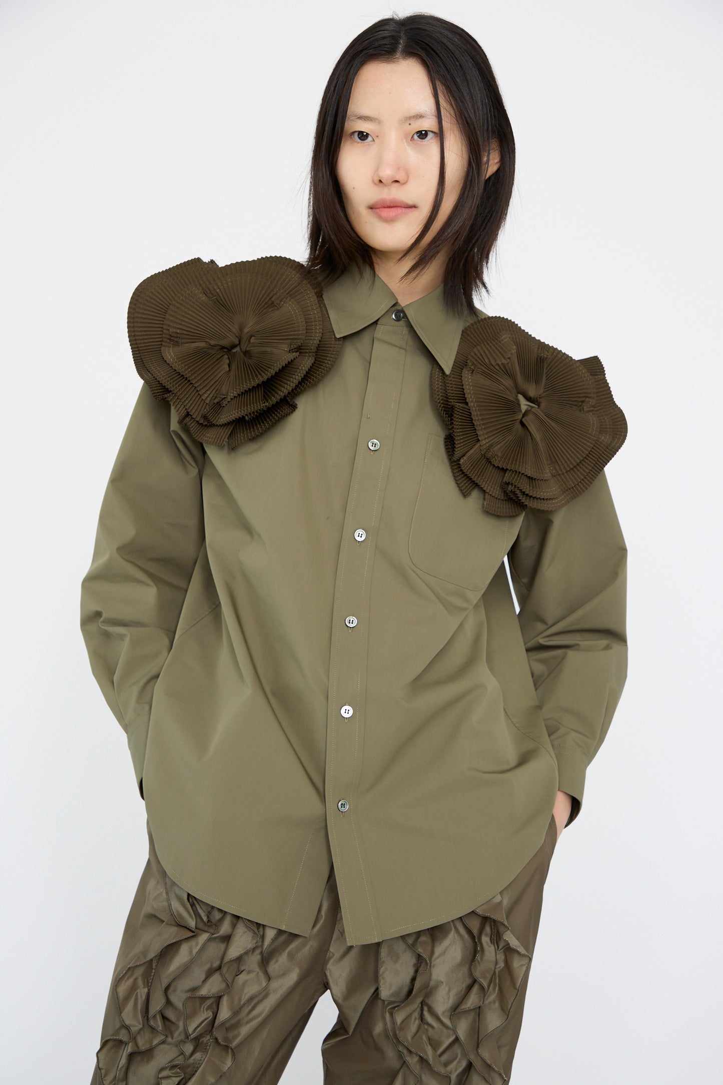 A model poses against a plain background wearing the TOGA ARCHIVES Flower Motif Shirt in Khaki, featuring an olive-green button-up design with large pleated and ruffled flower shapes on the shoulders, made from a soft cotton blend.