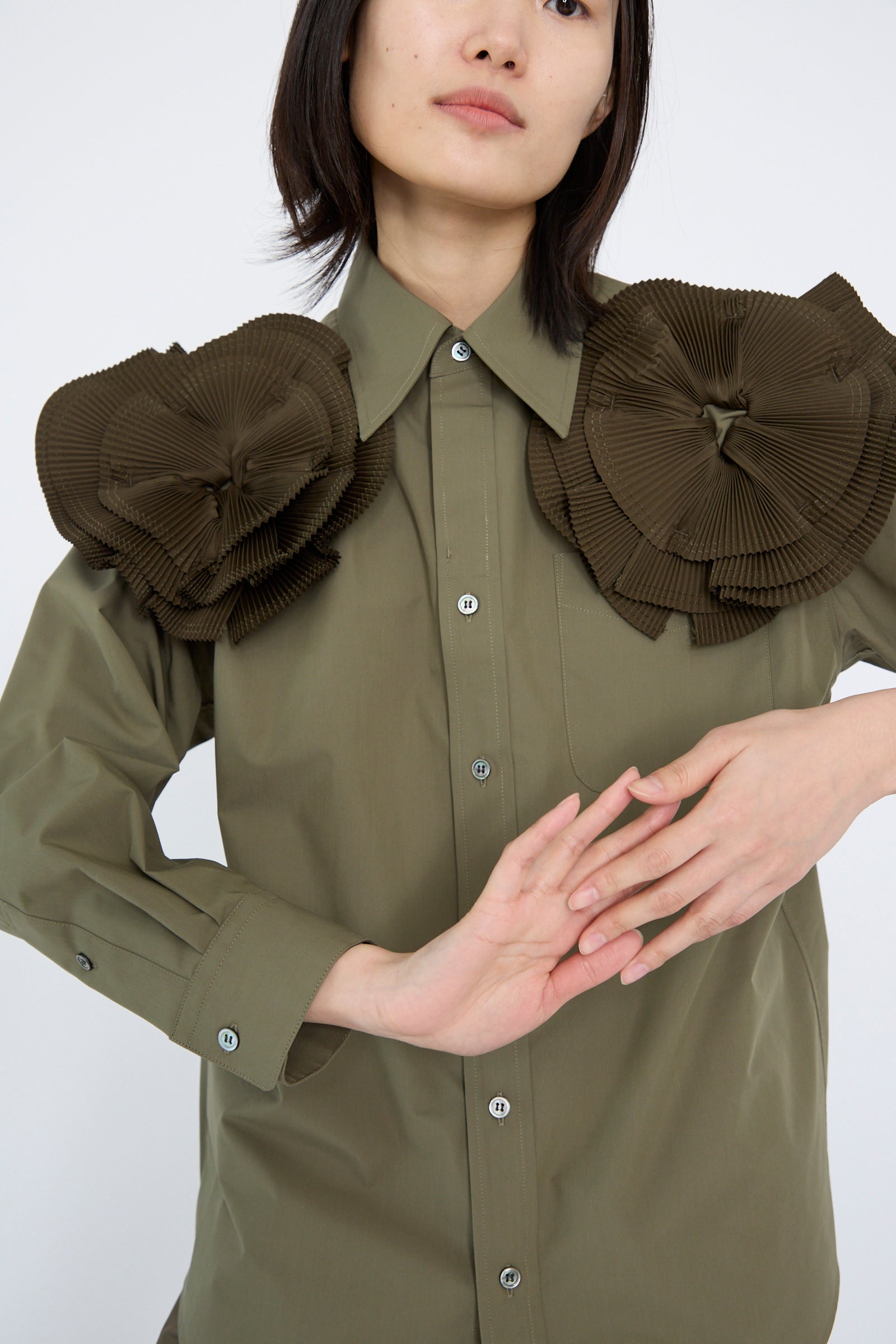 A person wearing a Flower Motif Shirt in Khaki by TOGA ARCHIVES, featuring a relaxed fit and cotton blend material, with ruffled flower shapes on the shoulders, holding their hands together.