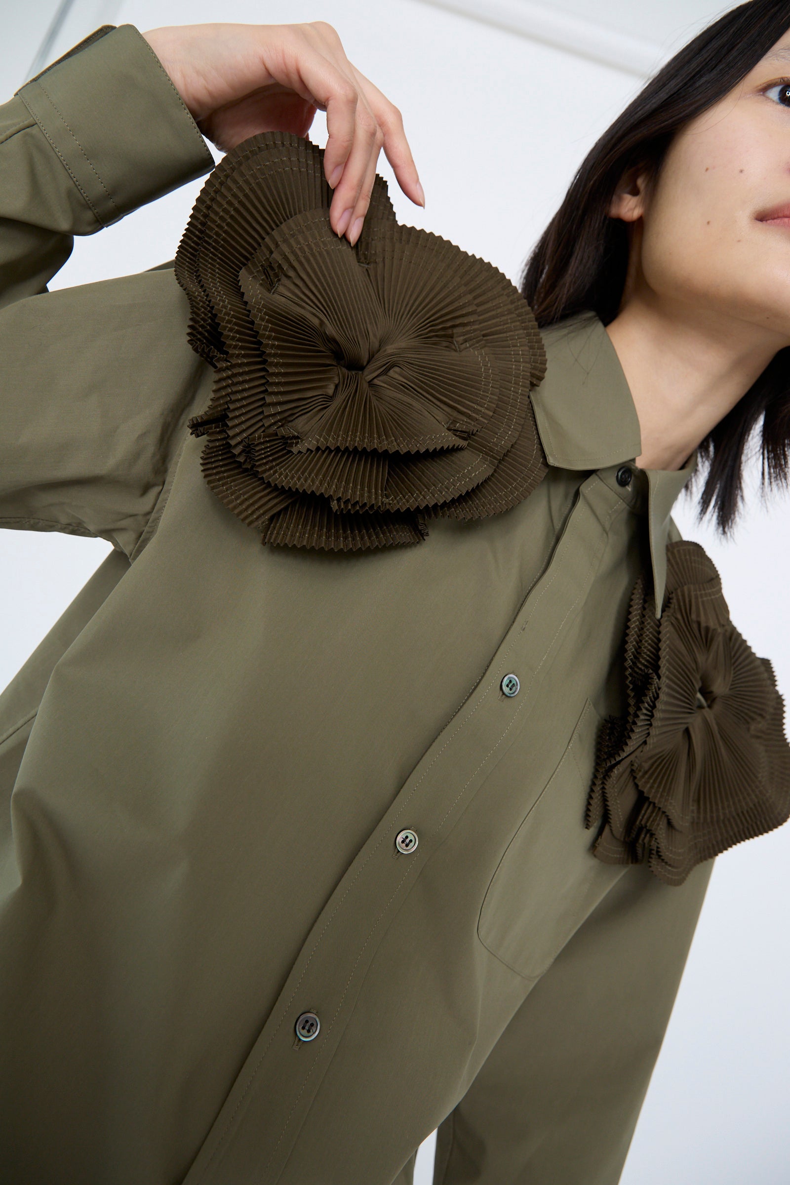 A person wearing the Flower Motif Shirt in Khaki by TOGA ARCHIVES, a green cotton blend button-up with a relaxed fit, featuring large pleated brown fabric embellishments resembling ruffled flower shapes on the shoulders.