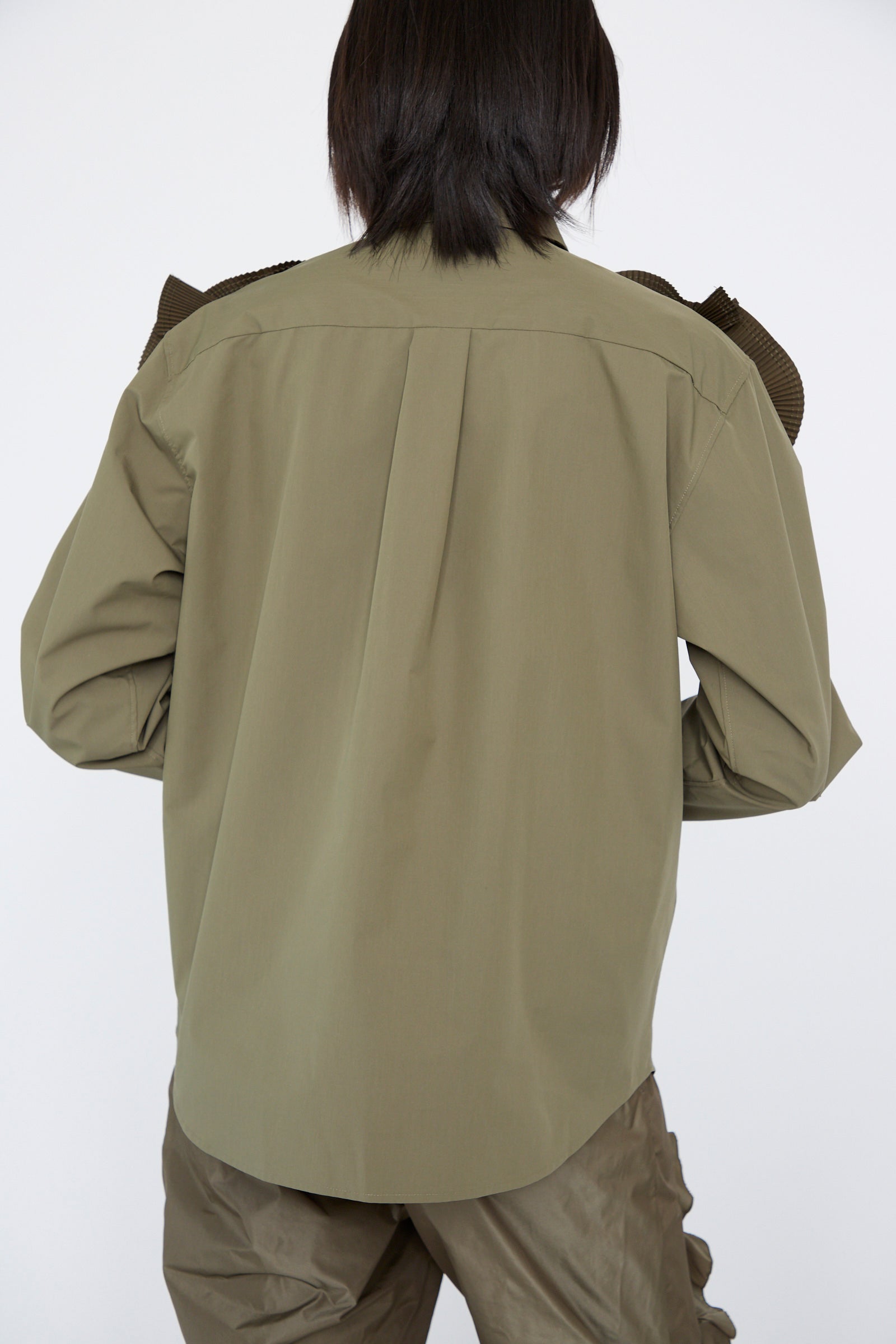 Someone with shoulder-length dark hair is wearing a TOGA ARCHIVES Flower Motif Shirt in khaki with relaxed fit, paired with brown pants, facing away from the camera.