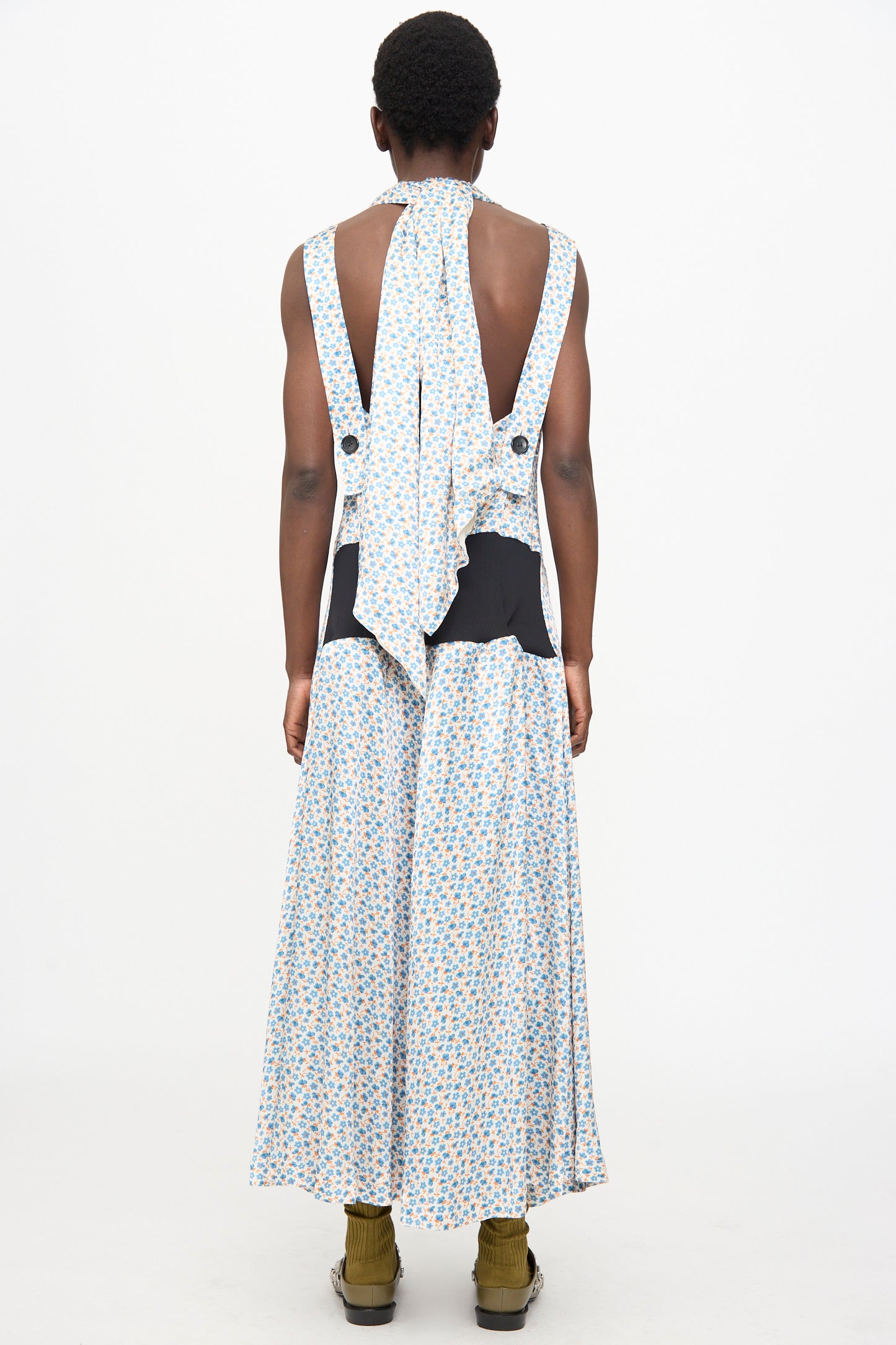 A person with short hair wears the TOGA ARCHIVES Flower Print Dress in White, a satin sleeveless maxi with black waist accents and a scarf-detail neck. 