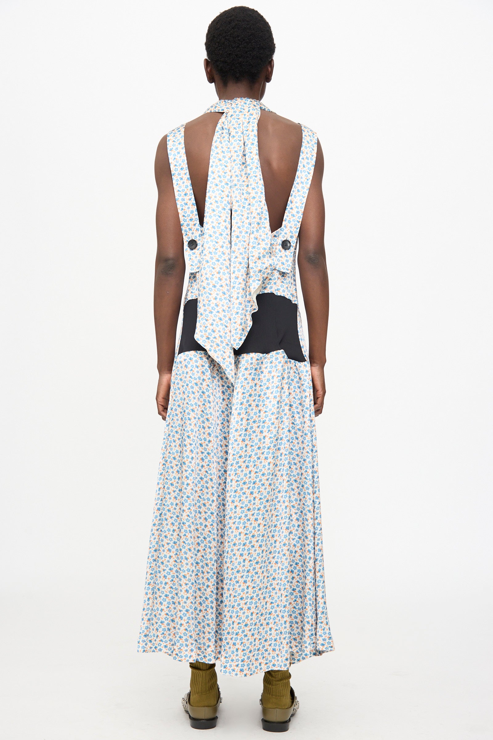 A person with short hair wears the TOGA ARCHIVES Flower Print Dress in White, a satin sleeveless maxi with black waist accents and a scarf-detail neck. 
