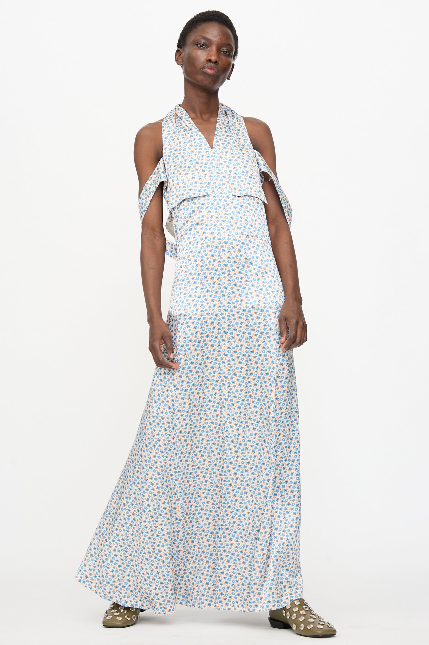 A person stands against a white background wearing the TOGA ARCHIVES Flower Print Dress in White, a sleeveless maxi dress made in Japan with blue and white floral prints and tie details, paired with olive green studded shoes.