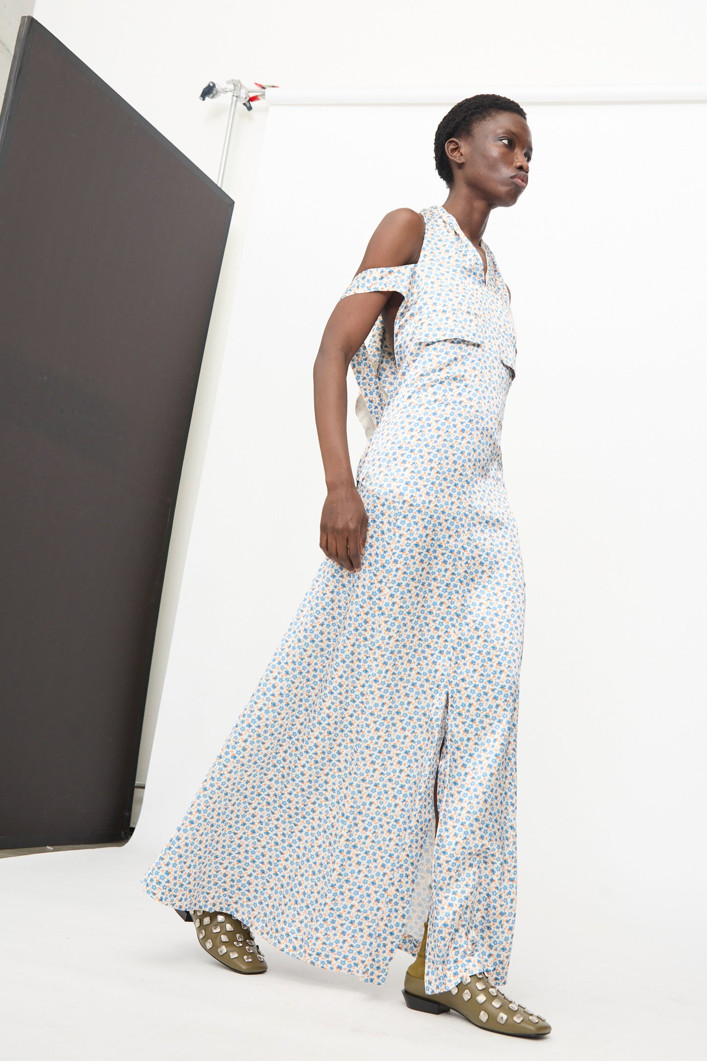 A person wearing the TOGA ARCHIVES Flower Print Dress in White and olive green shoes with white spots walks in a studio with a plain background.
