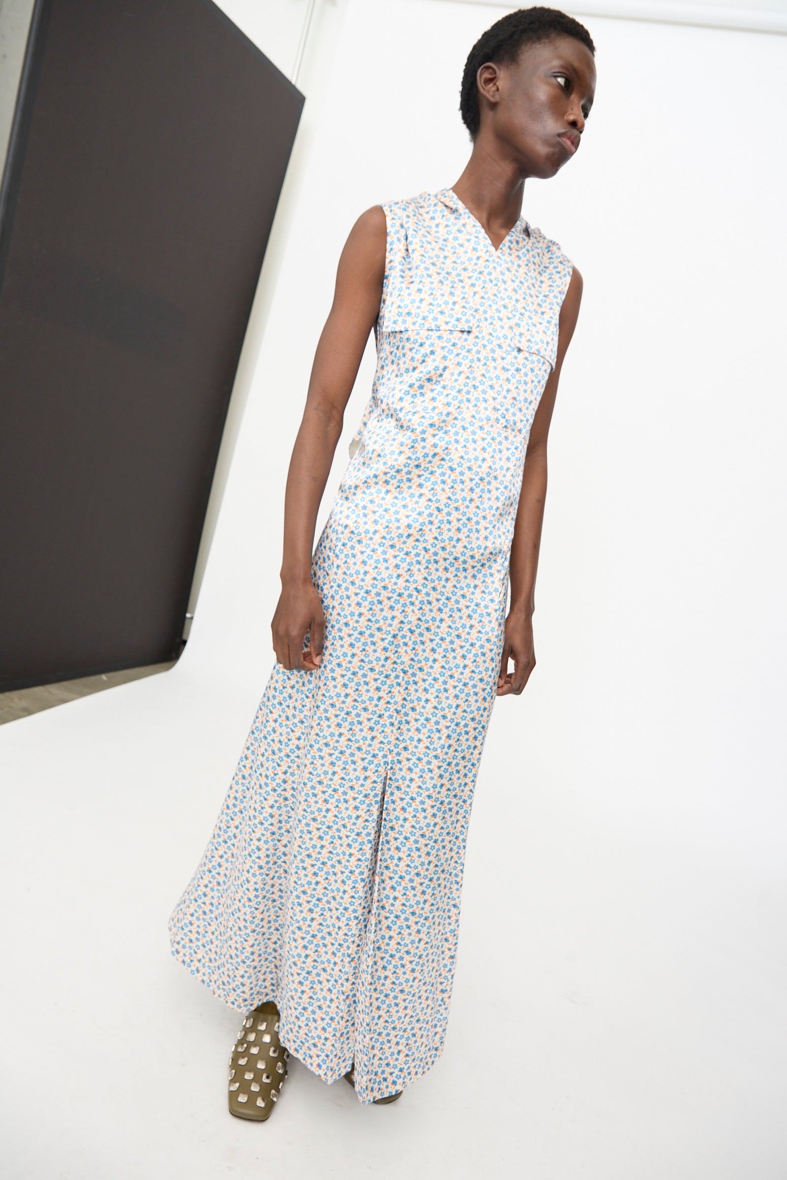 A person stands in a minimalist studio wearing the TOGA ARCHIVES Flower Print Dress in White, a sleeveless maxi made in Japan with small blue and yellow motifs, paired with brown shoes featuring white spots.