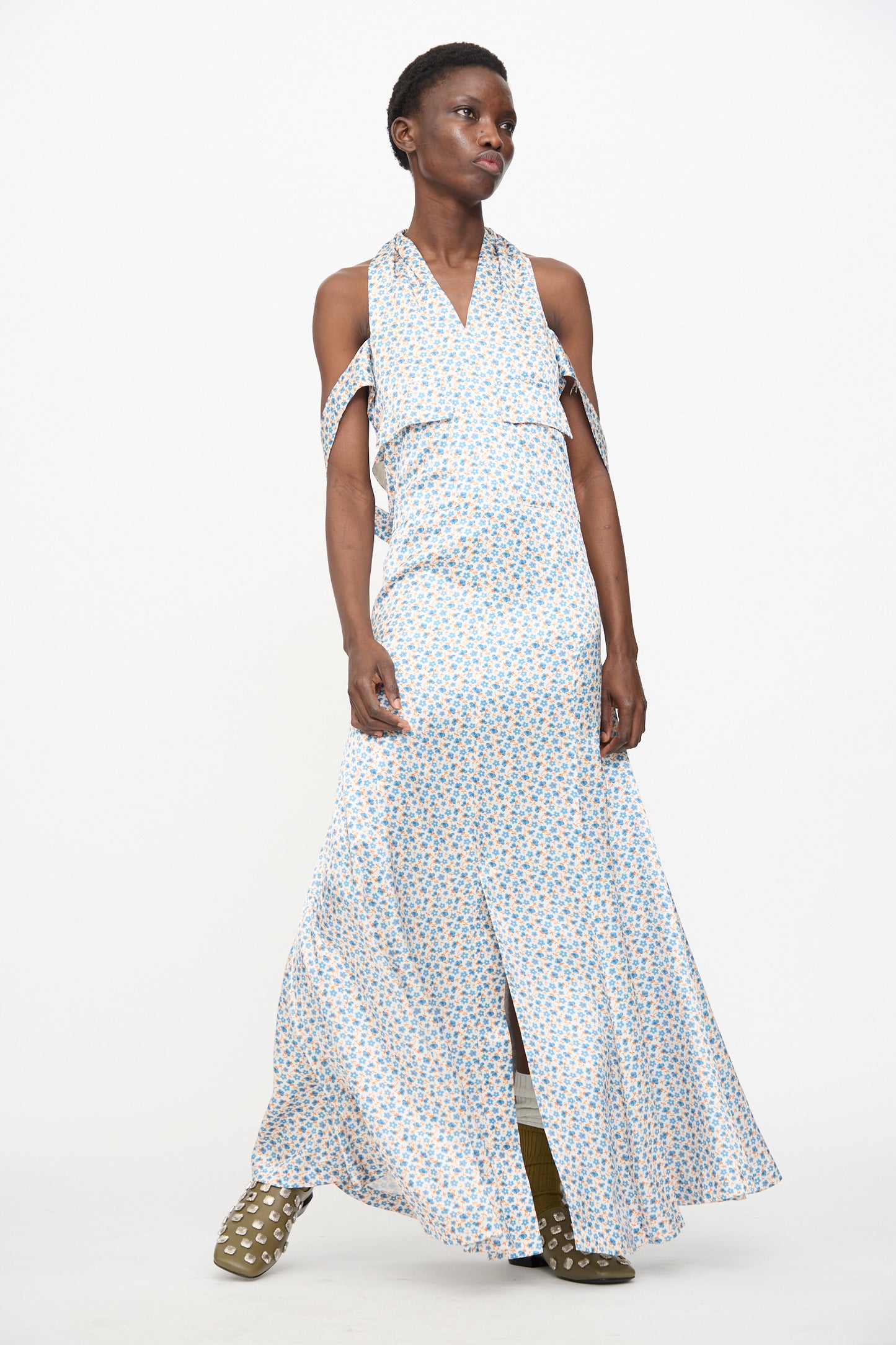 A person stands against a white background wearing the TOGA ARCHIVES Flower Print Dress in White, a long, sleeveless maxi made in Japan with light blue floral satin and shoulder straps, paired with olive green ankle boots with white spots.