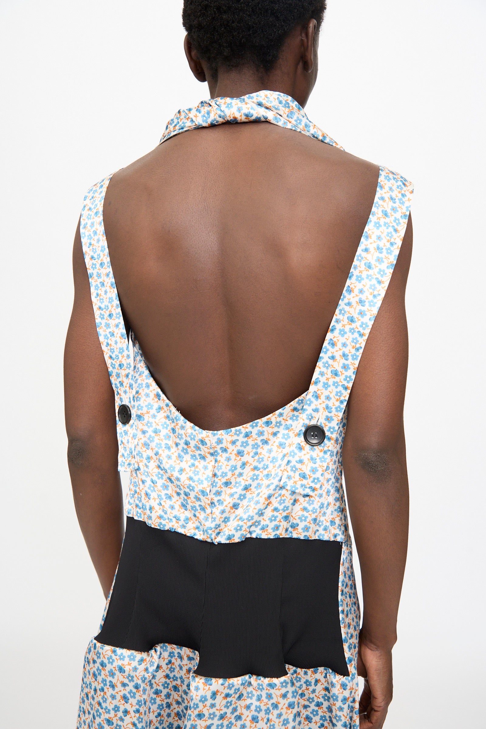 A person is shown from behind wearing the TOGA ARCHIVES Flower Print Dress in White, a sleeveless Japanese maxi dress with blue and orange floral satin, black waist panel, and large buttons, set against a plain background.