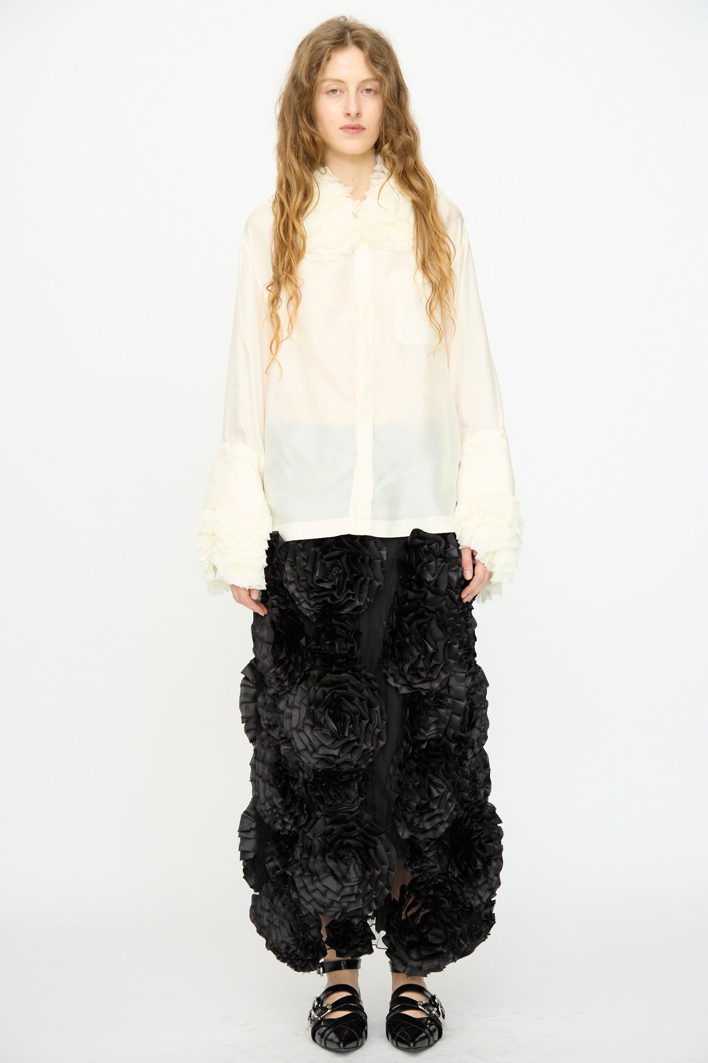 A person stands against a plain white background wearing the TOGA ARCHIVES Frill Embroidery Blouse in Off White, featuring textured cuffs, paired with a black skirt embellished with large 3D floral details.