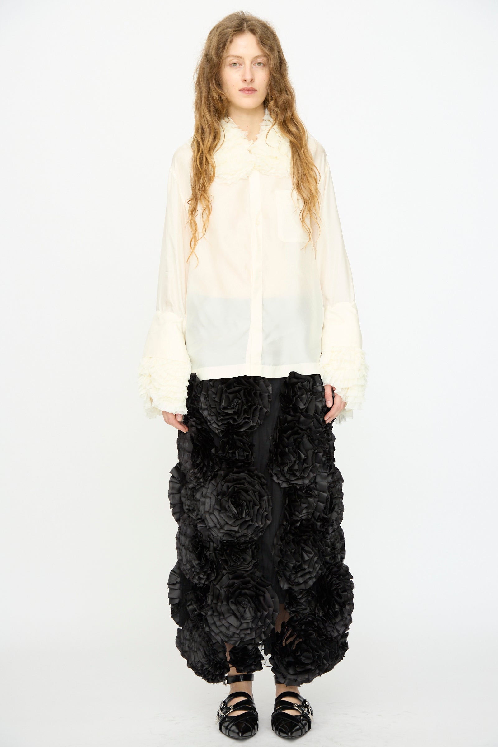 A person stands against a plain white background wearing the TOGA ARCHIVES Frill Embroidery Blouse in Off White, featuring textured cuffs, paired with a black skirt embellished with large 3D floral details.