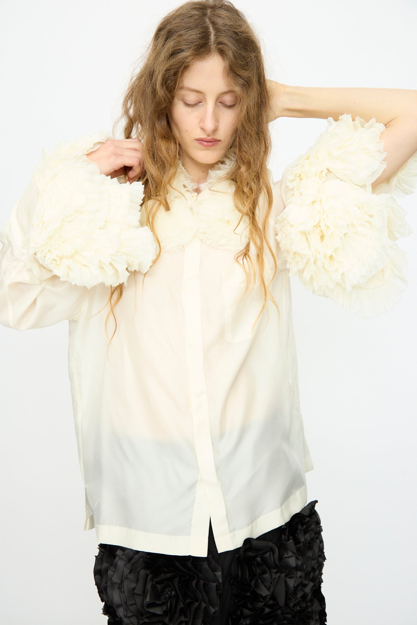 A person with long wavy hair adjusts the TOGA ARCHIVES Frill Embroidery Blouse in Off White, featuring sheer fabric and large ruffled sleeves, paired with black textured shorts against a plain white backdrop.