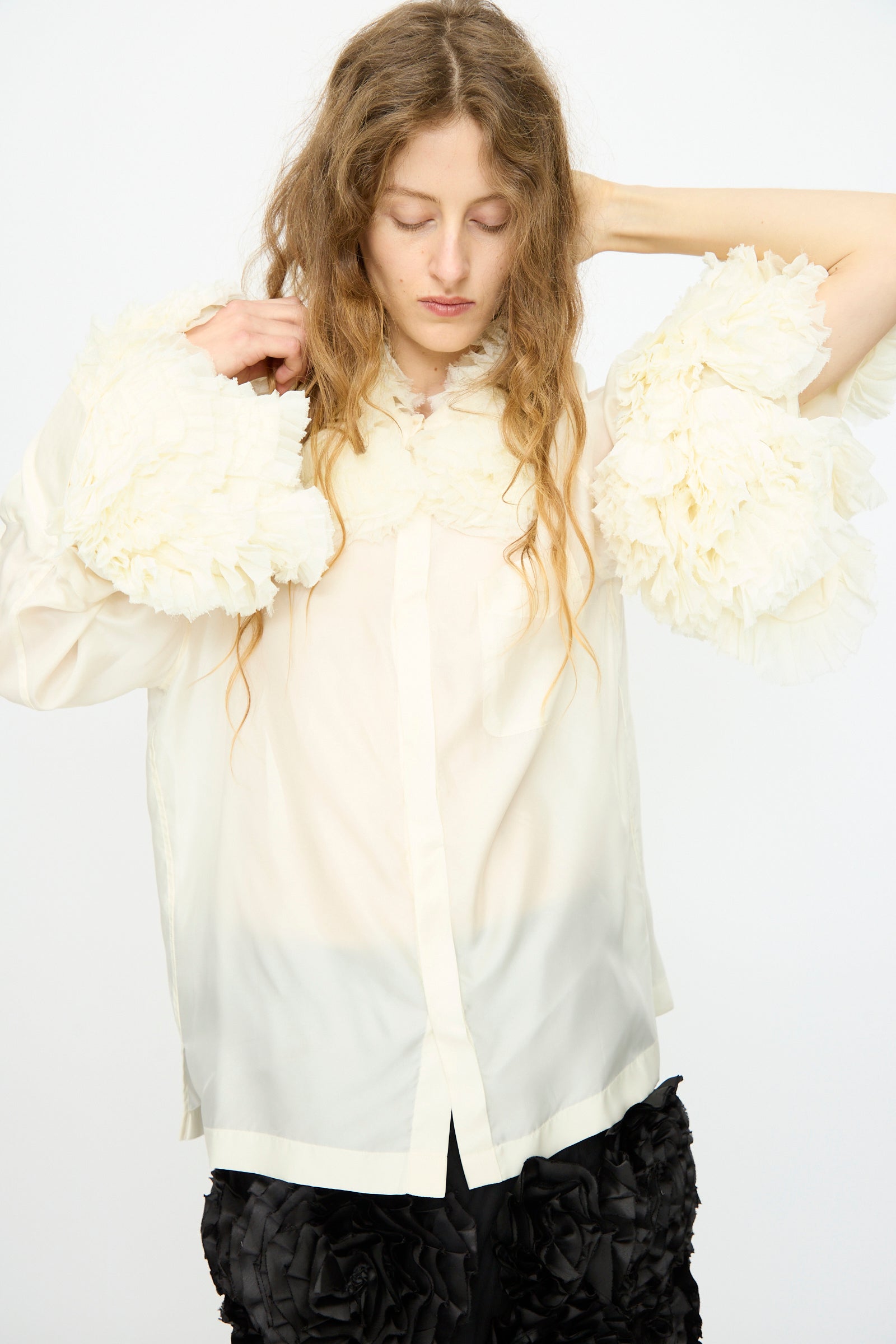 A person with long wavy hair adjusts the TOGA ARCHIVES Frill Embroidery Blouse in Off White, featuring sheer fabric and large ruffled sleeves, paired with black textured shorts against a plain white backdrop.