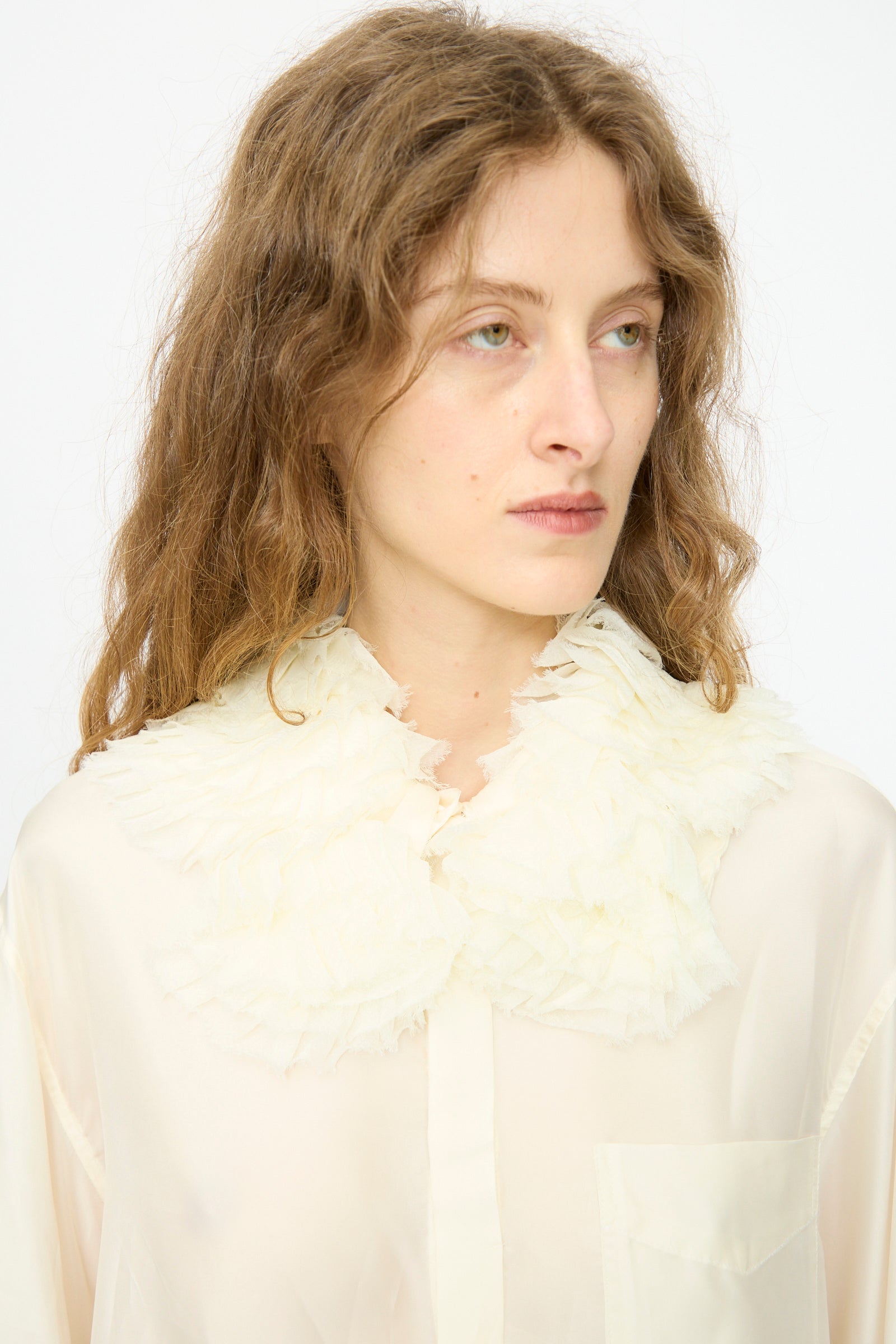 A person with wavy light brown hair wears the Frill Embroidery Blouse in Off White by TOGA ARCHIVES, featuring a sheer fabric and large textured detachable collar, posing against a plain white background.