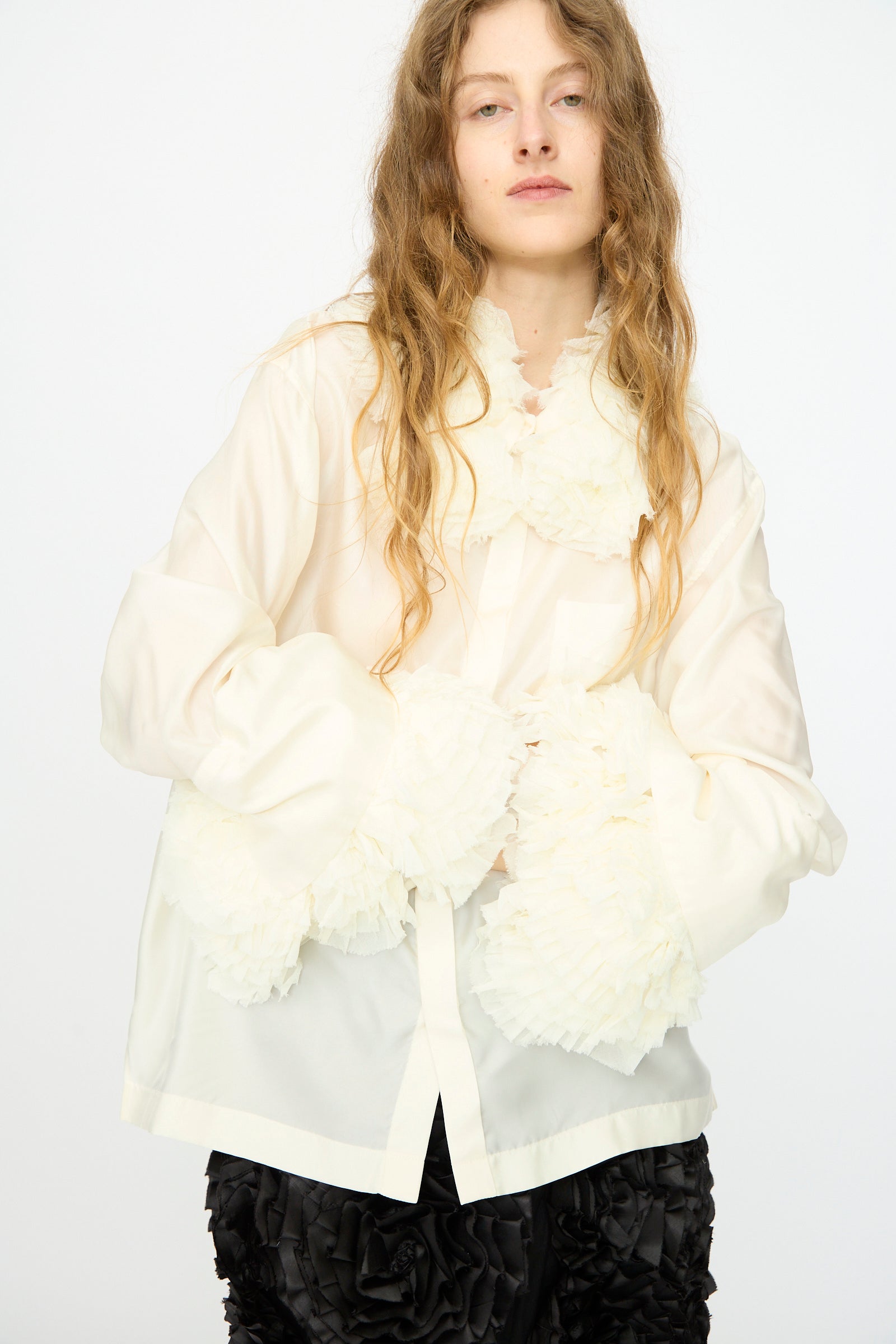 A person wears the TOGA ARCHIVES Frill Embroidery Blouse in Off White, featuring ruffled details and a detachable frill collar, paired with black textured pants, standing against a plain white background.