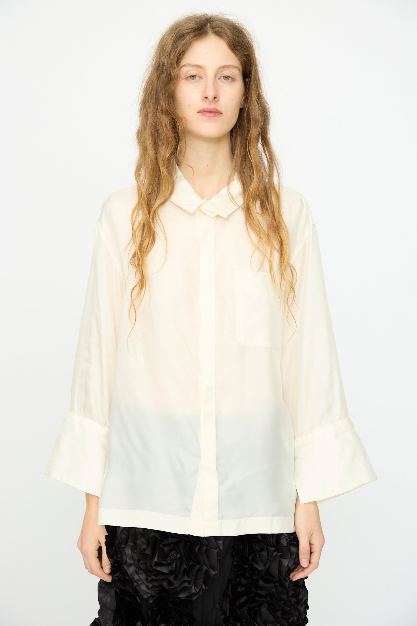A person with long wavy hair wears the TOGA ARCHIVES Frill Embroidery Blouse in Off White and black textured pants, standing against a plain white background.