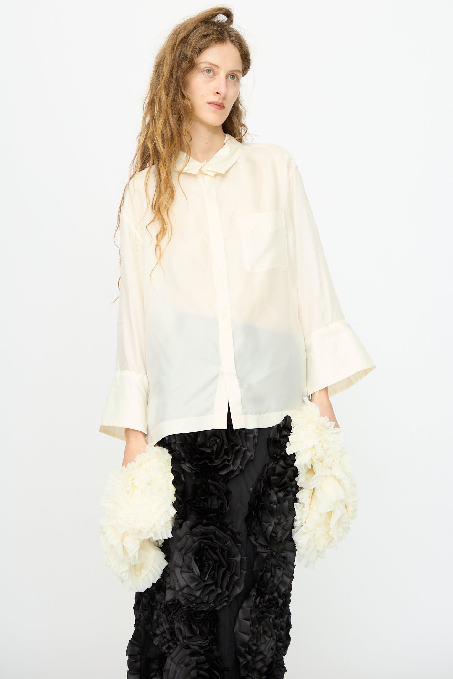 A woman with long wavy hair wears the TOGA ARCHIVES Frill Embroidery Blouse in Off White and a black skirt with large fabric flowers, standing against a plain white background.