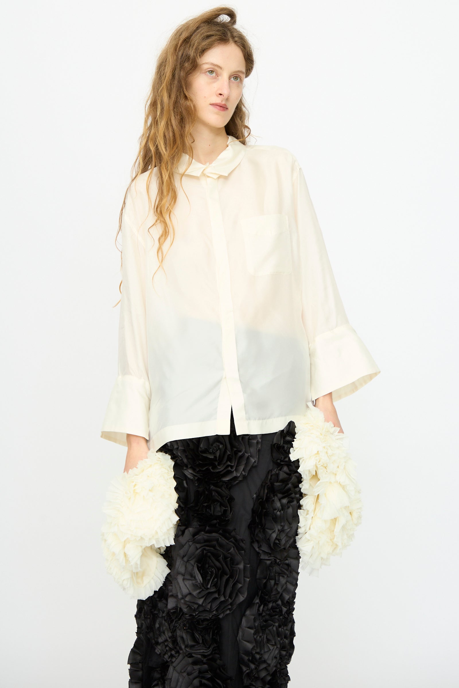 A woman with long wavy hair wears the TOGA ARCHIVES Frill Embroidery Blouse in Off White and a black skirt with large fabric flowers, standing against a plain white background.