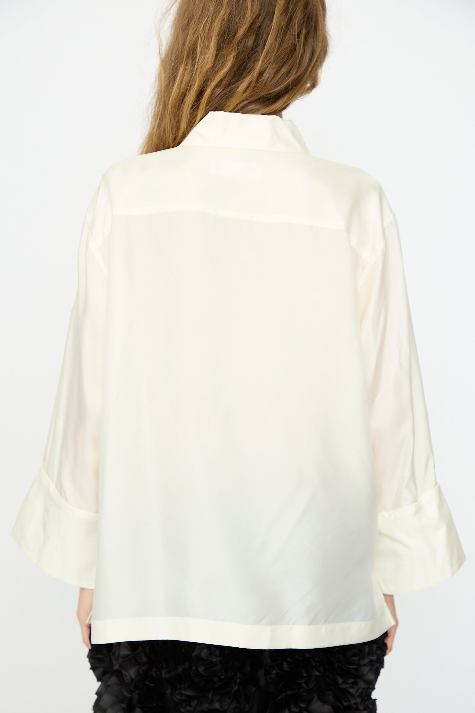 A person with long, light brown hair is seen from the back wearing the TOGA ARCHIVES Frill Embroidery Blouse in Off White and a black textured skirt against a plain background.