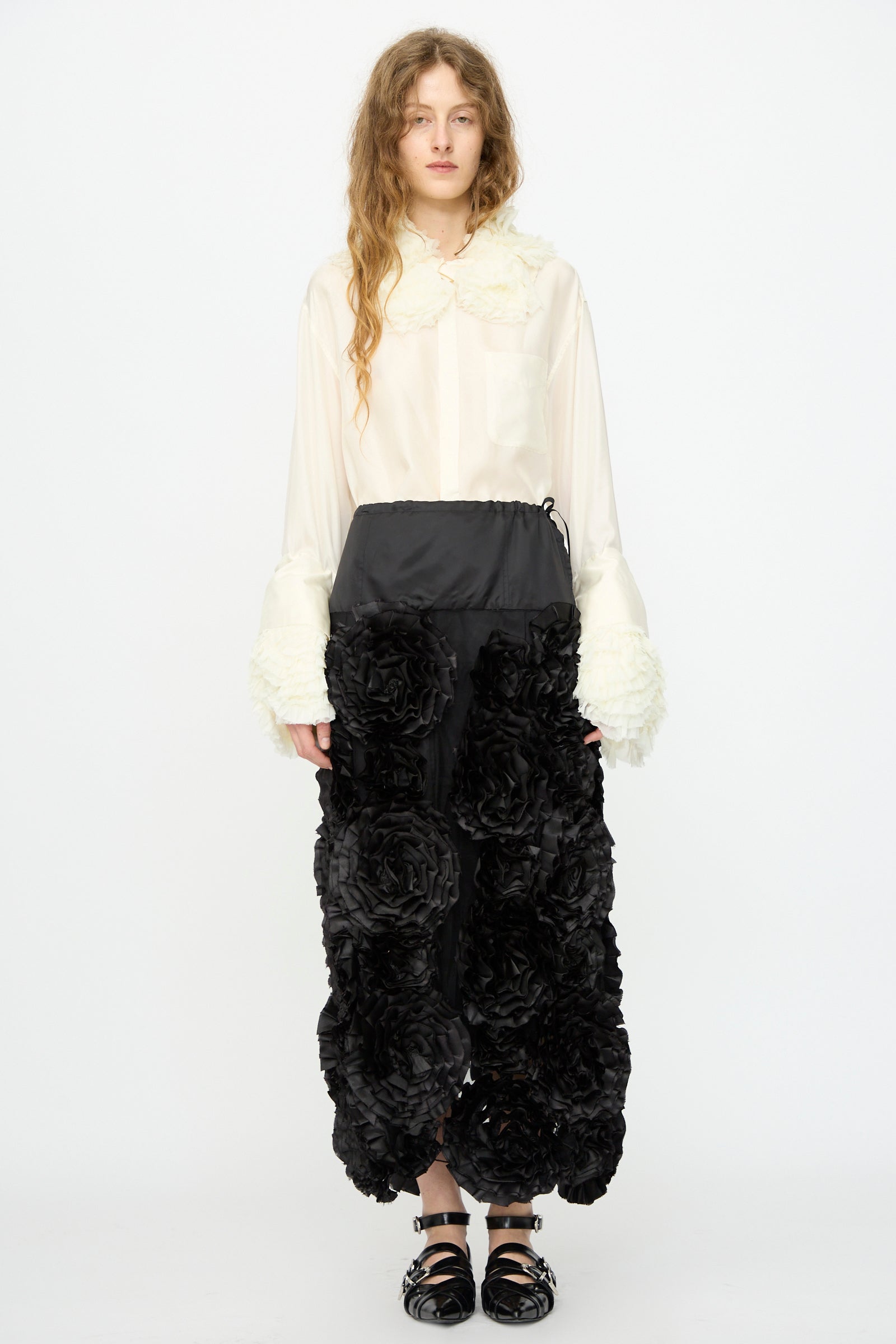 A person stands facing forward wearing a sheer white blouse with textured cuffs and collar, paired with the TOGA ARCHIVES Frill Embroidery Skirt in Black and black sandals.