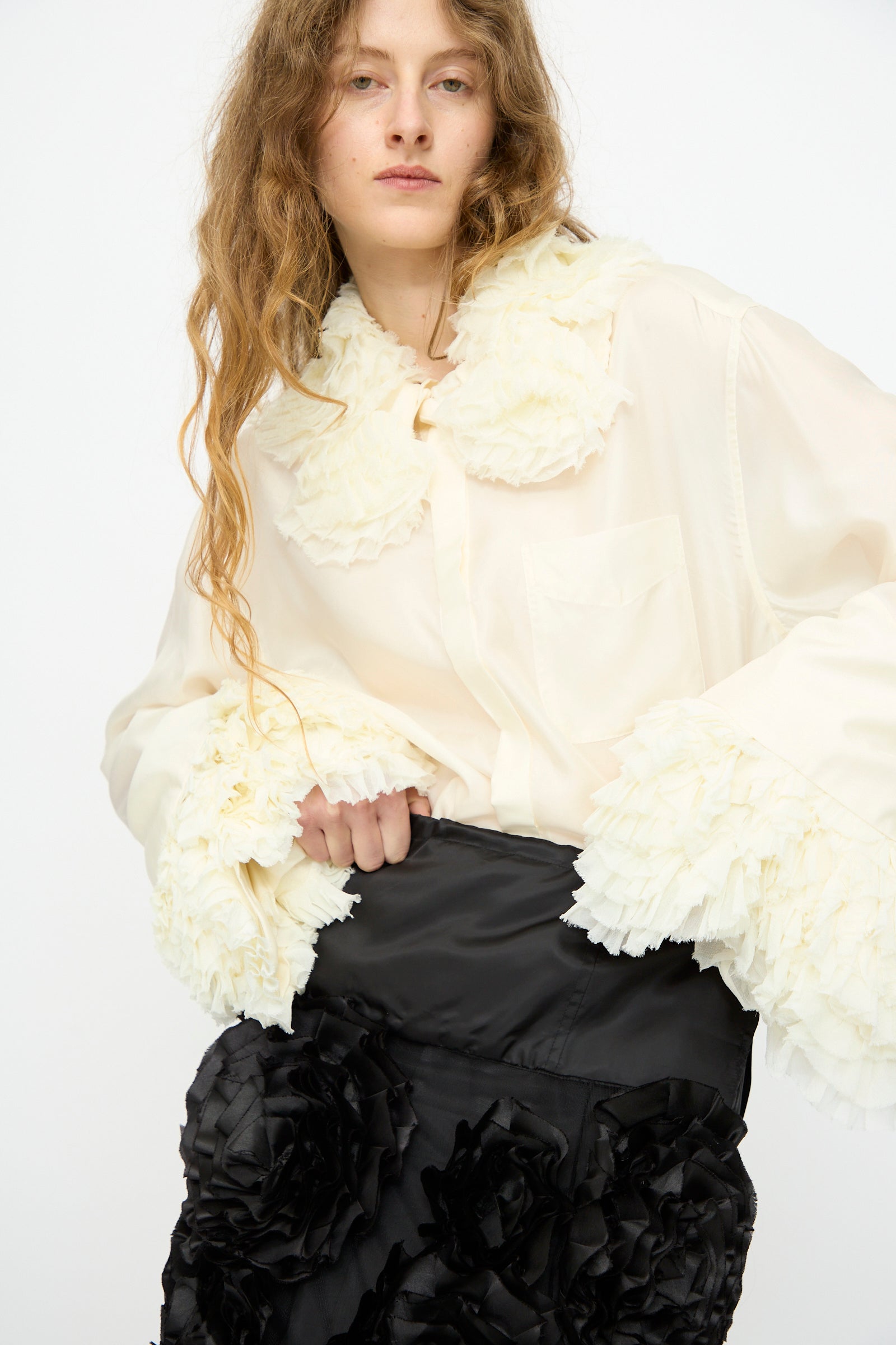 A model in a cream blouse with ruffled collar and cuffs pairs it with TOGA ARCHIVES’ Frill Embroidery Skirt in Black, featuring large textured florals, against a plain white background.