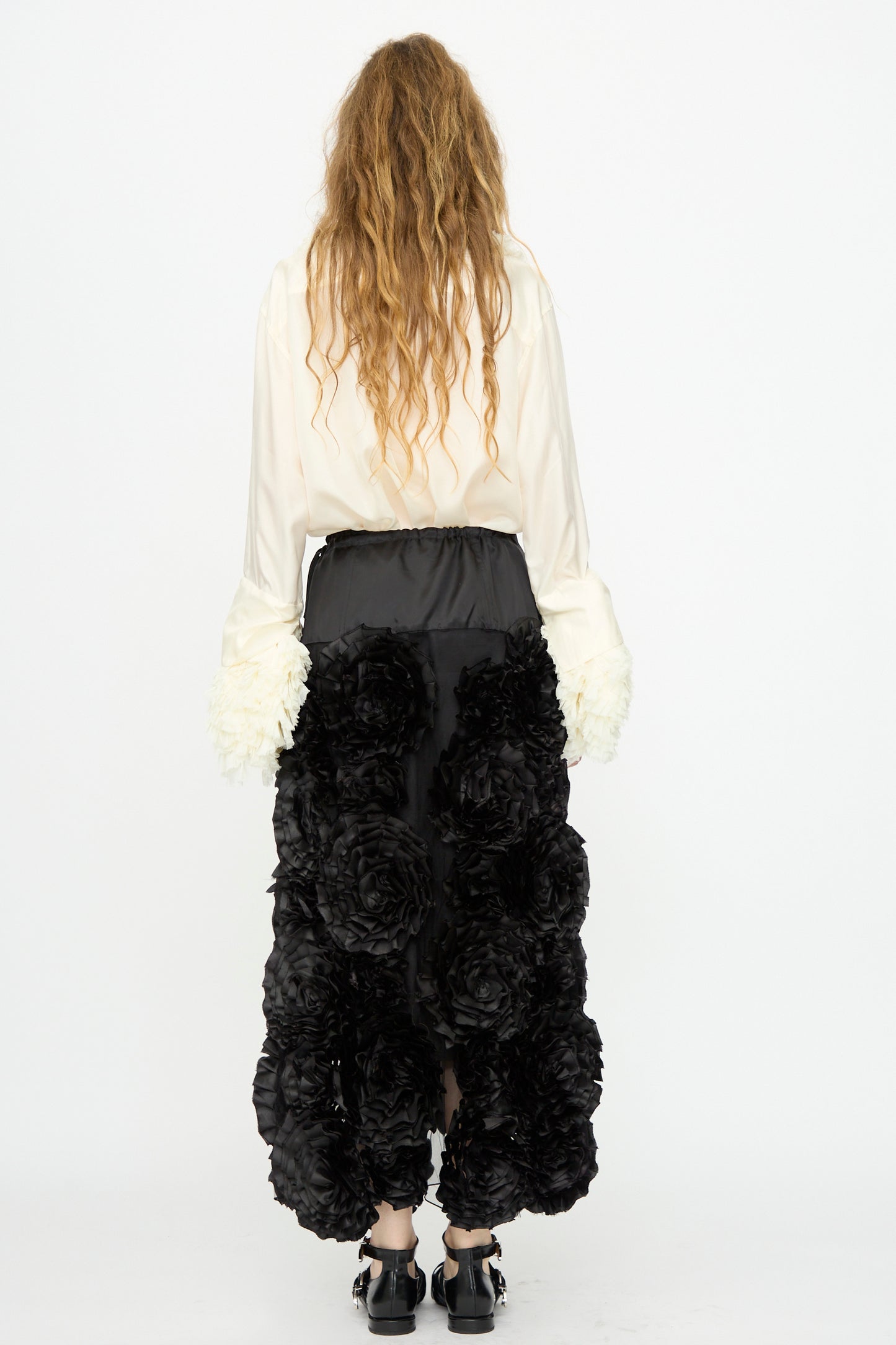 A person with long wavy hair faces away, wearing a cream blouse with textured white cuffs and the TOGA ARCHIVES Frill Embroidery Skirt in Black.