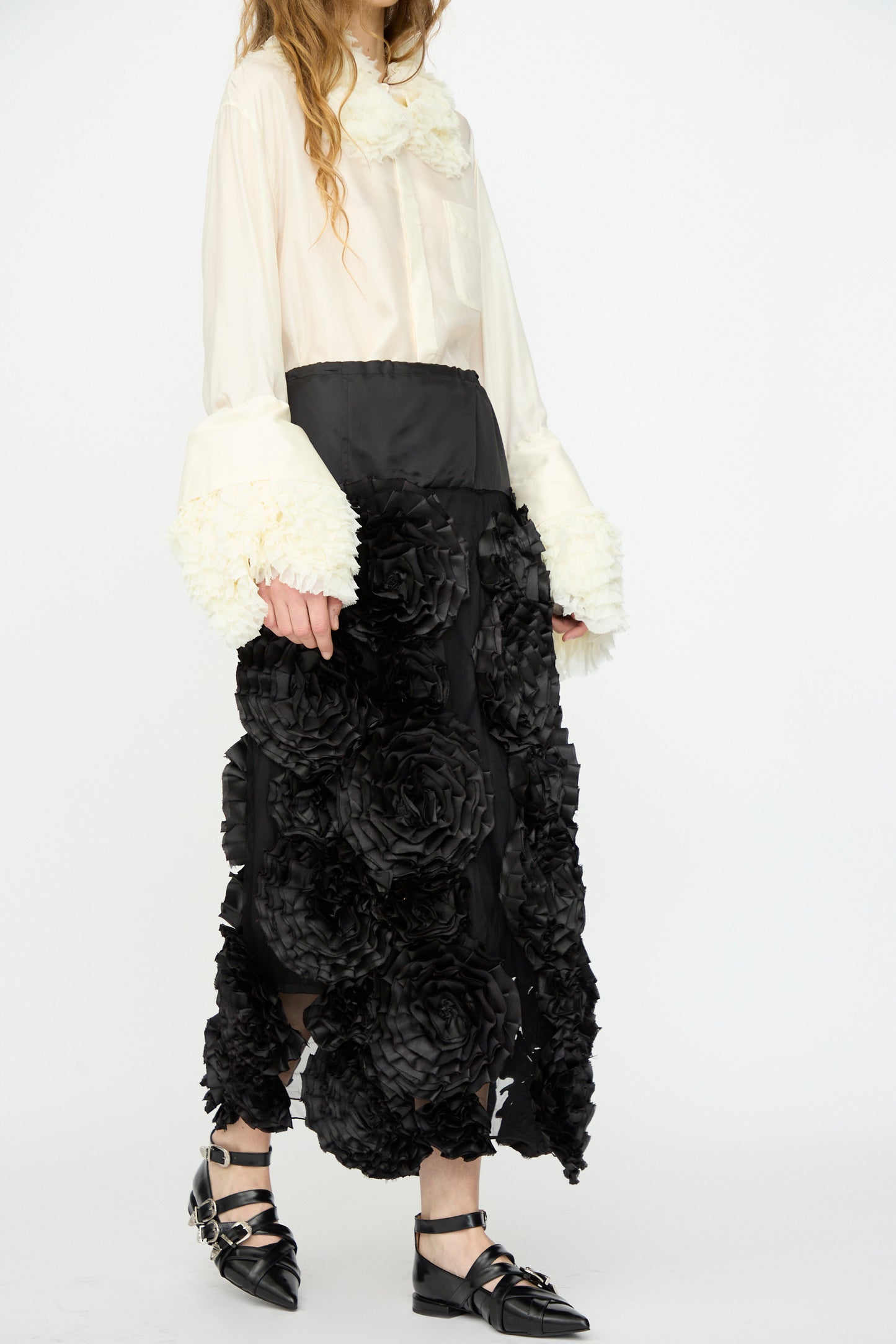 A person wearing a white blouse with textured cuffs, the TOGA ARCHIVES Frill Embroidery Skirt in Black, and black pointed strap shoes.