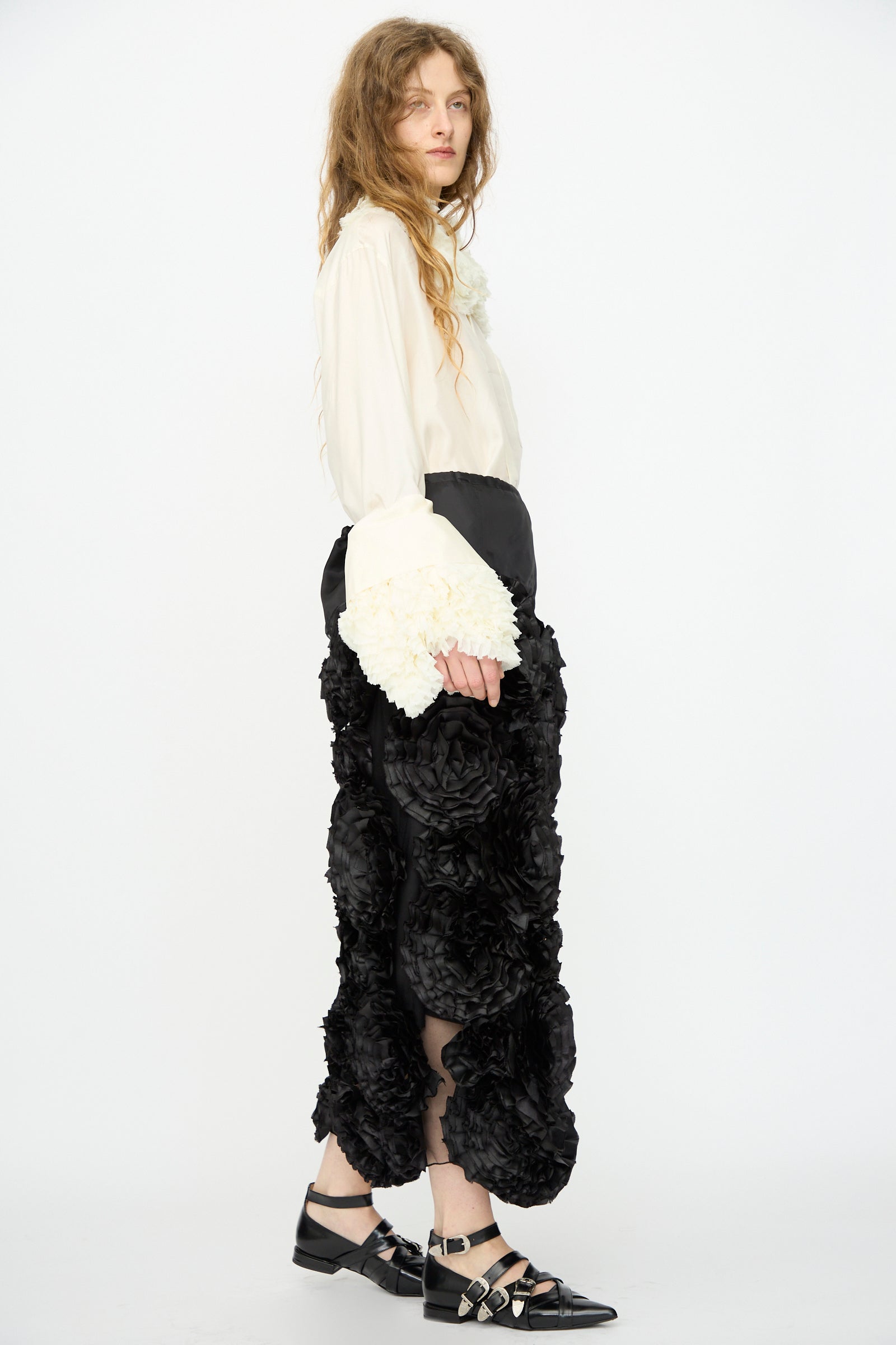 A person wears the TOGA ARCHIVES Frill Embroidery Skirt in Black with a cream long-sleeve blouse featuring voluminous cuffs and black strappy shoes, standing against a white background.