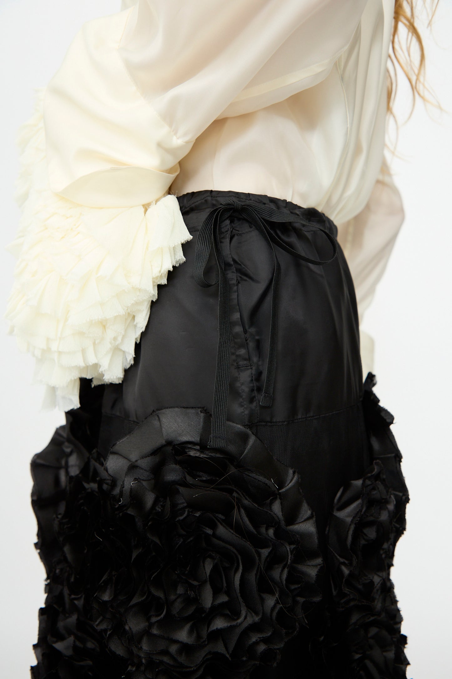 A person wearing a cream ruffled blouse and the TOGA ARCHIVES Frill Embroidery Skirt in Black, featuring large textured floral appliqués, shown from the side.