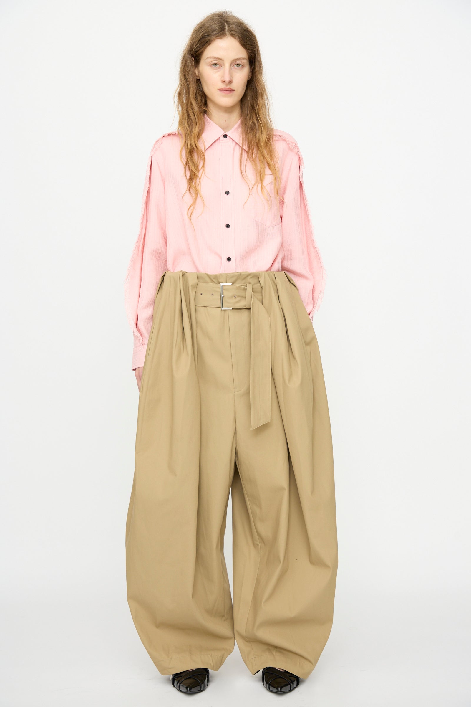 A person stands against a plain background wearing TOGA ARCHIVES High Twist Twill Wide Pants in Beige—high-waisted, wide-leg pants crafted from a soft cotton blend and finished with a belt for a polished look.