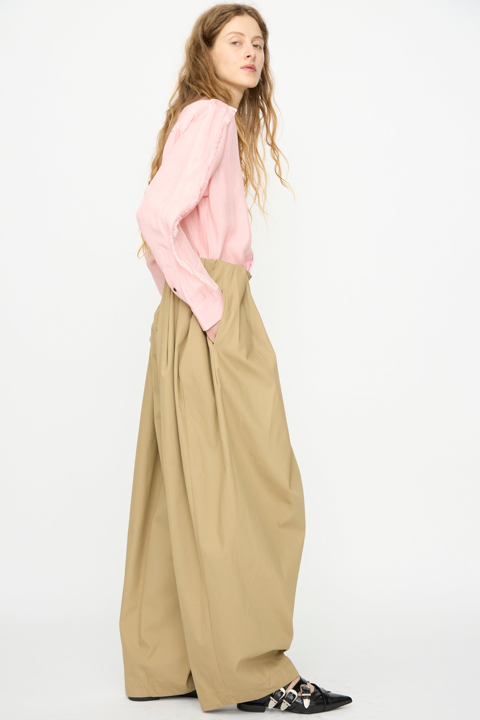 A person stands sideways against a plain white background, wearing TOGA ARCHIVES High Twist Twill Wide Pants in Beige, a pink long-sleeve shirt, and black pointed shoes.