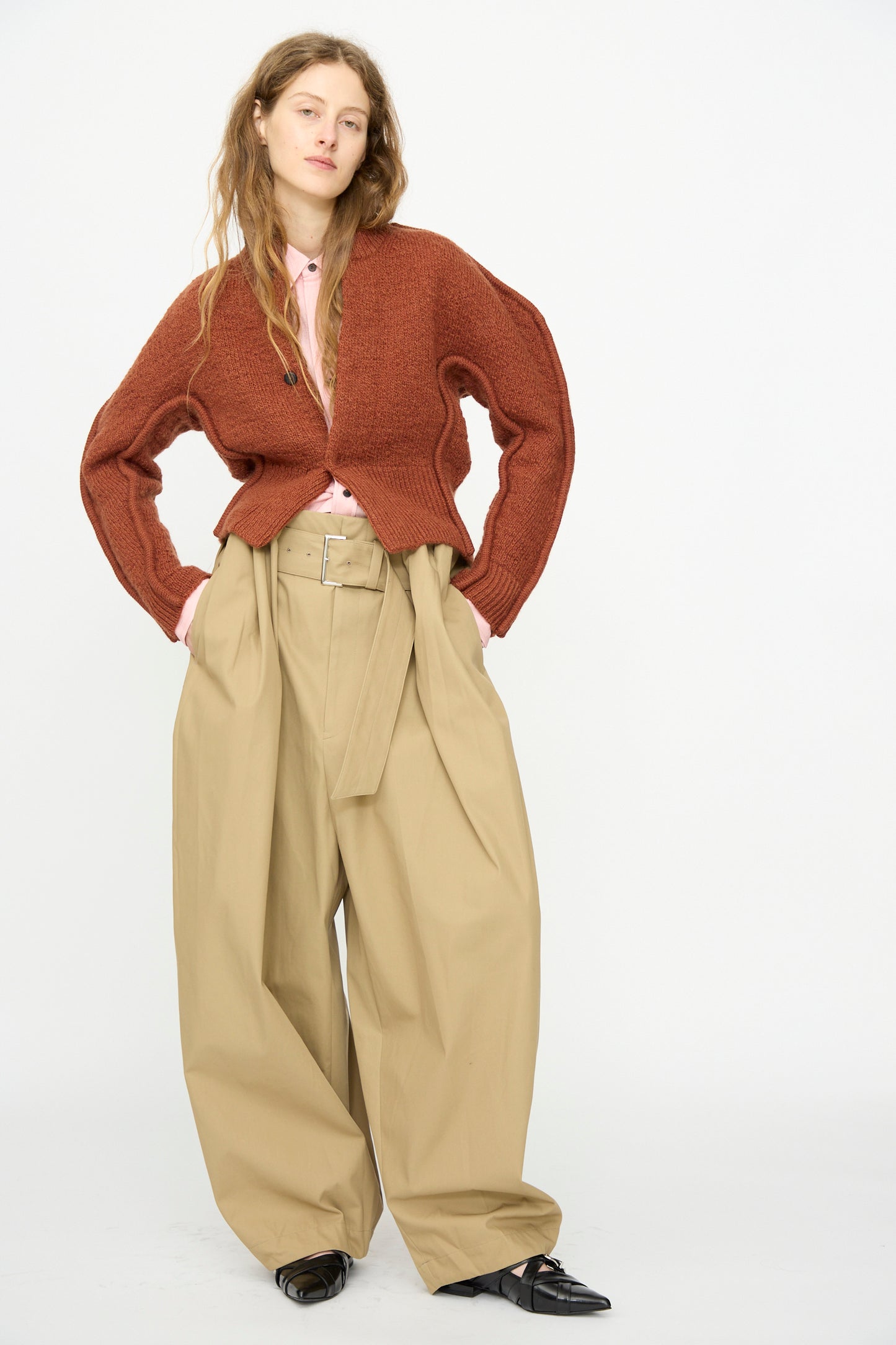 A person stands against a white background wearing TOGA ARCHIVES High Twist Twill Wide Pants in Beige, paired with a brown cardigan, pink shirt, and black shoes, hands on hips.