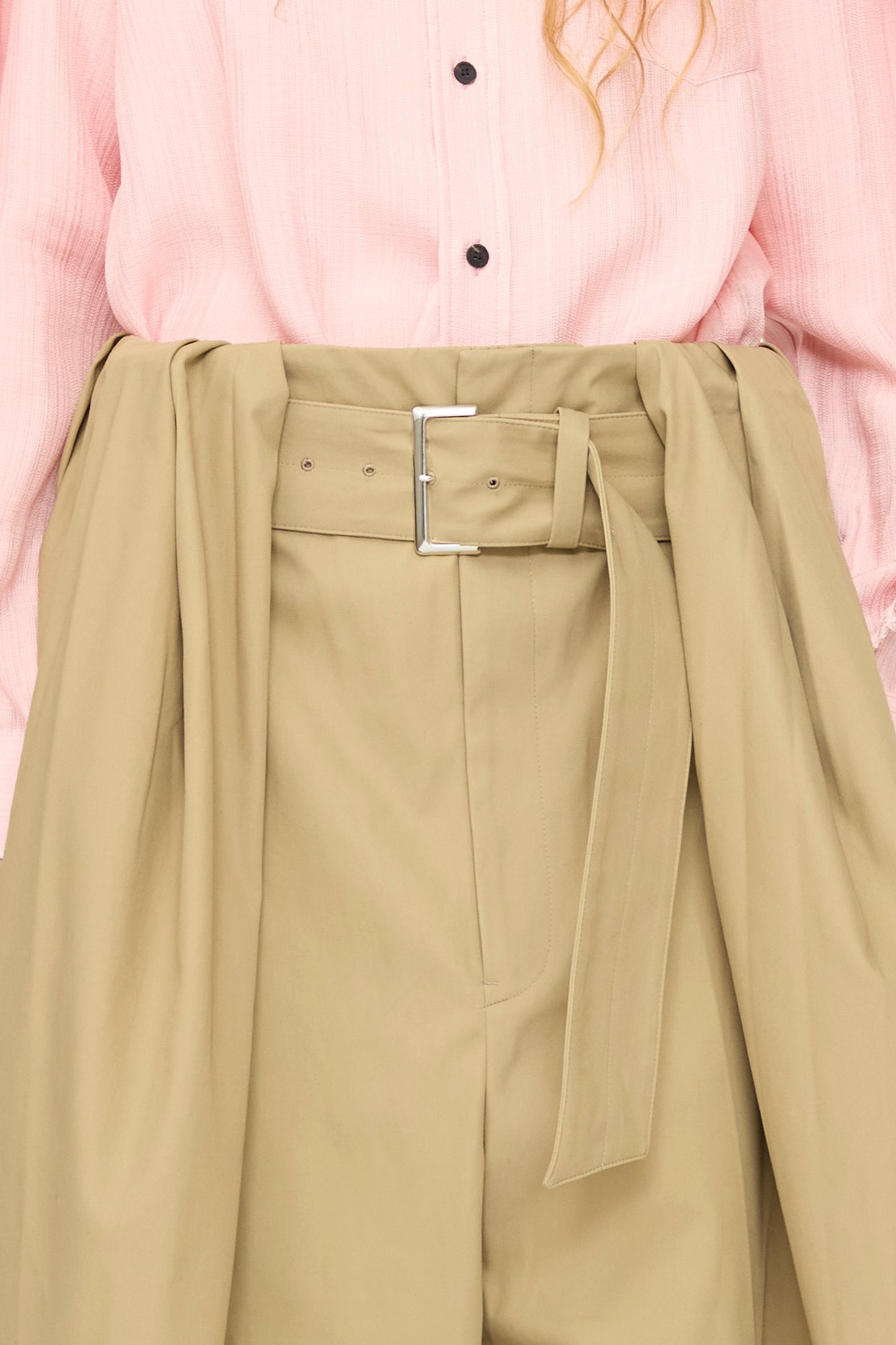A person wears TOGA ARCHIVES High Twist Twill Wide Pants in Beige, styled high-waisted with a belt and long fabric ties, paired with a tucked-in light pink button-up shirt.