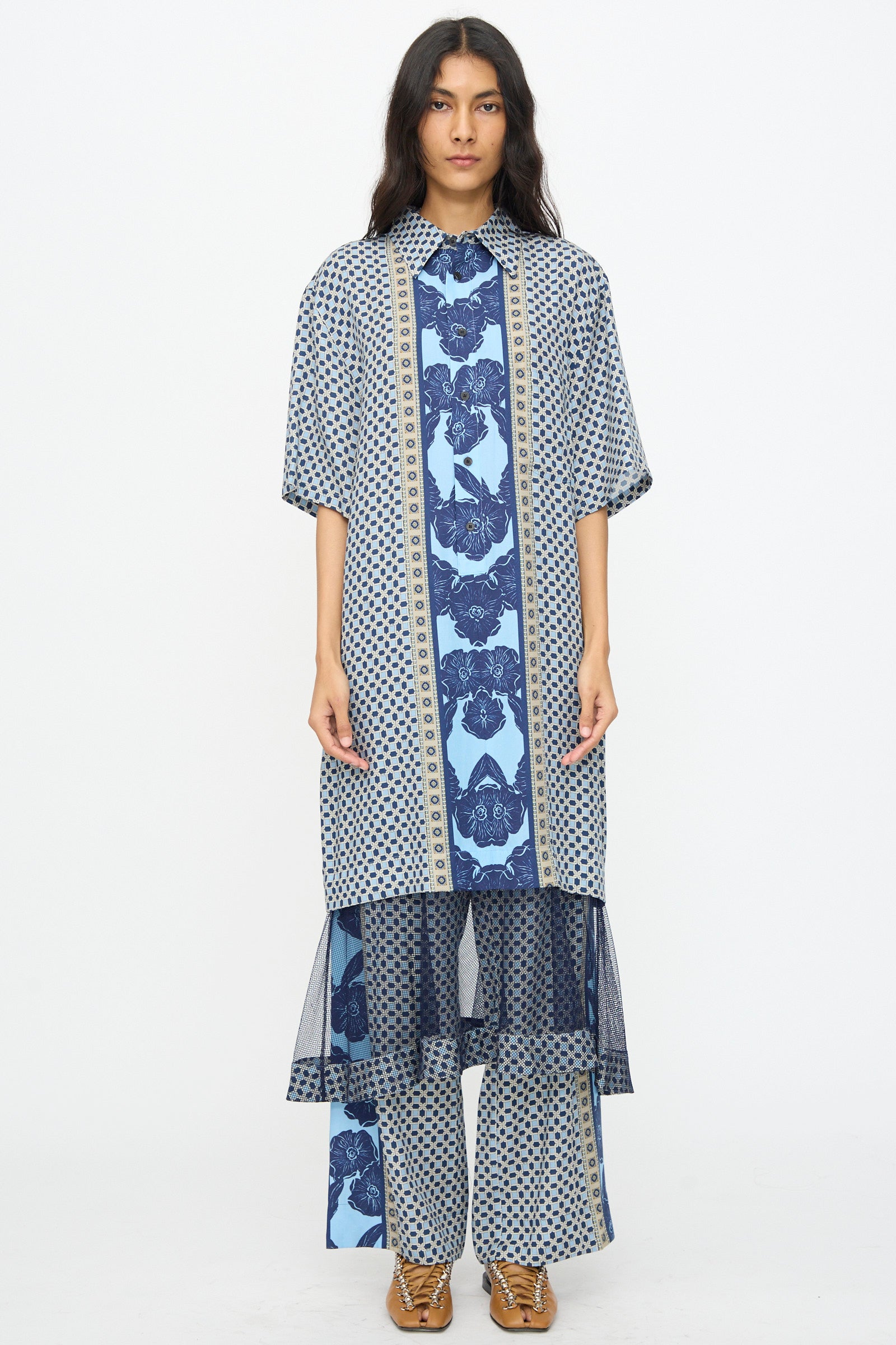 A woman wears the TOGA ARCHIVES Inner Print Dress in Light Blue, featuring floral and geometric patterns, styled with brown shoes against a plain white background.