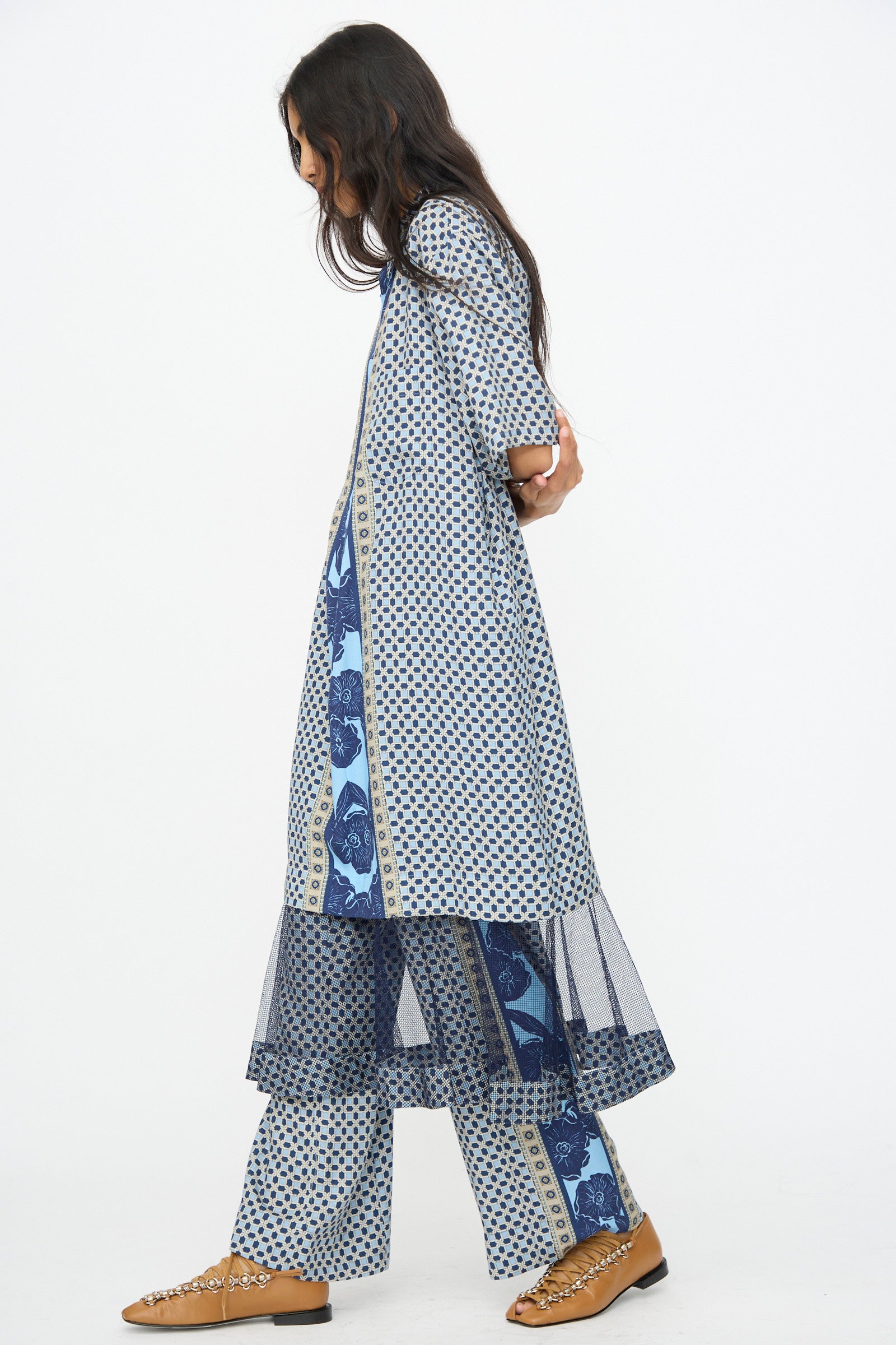 A woman in the TOGA ARCHIVES Inner Print Dress in Light Blue stands sideways against a white background, her look finished with long dark hair, brown flats, and a touch of elegance from the dress’s pleated skirt design.