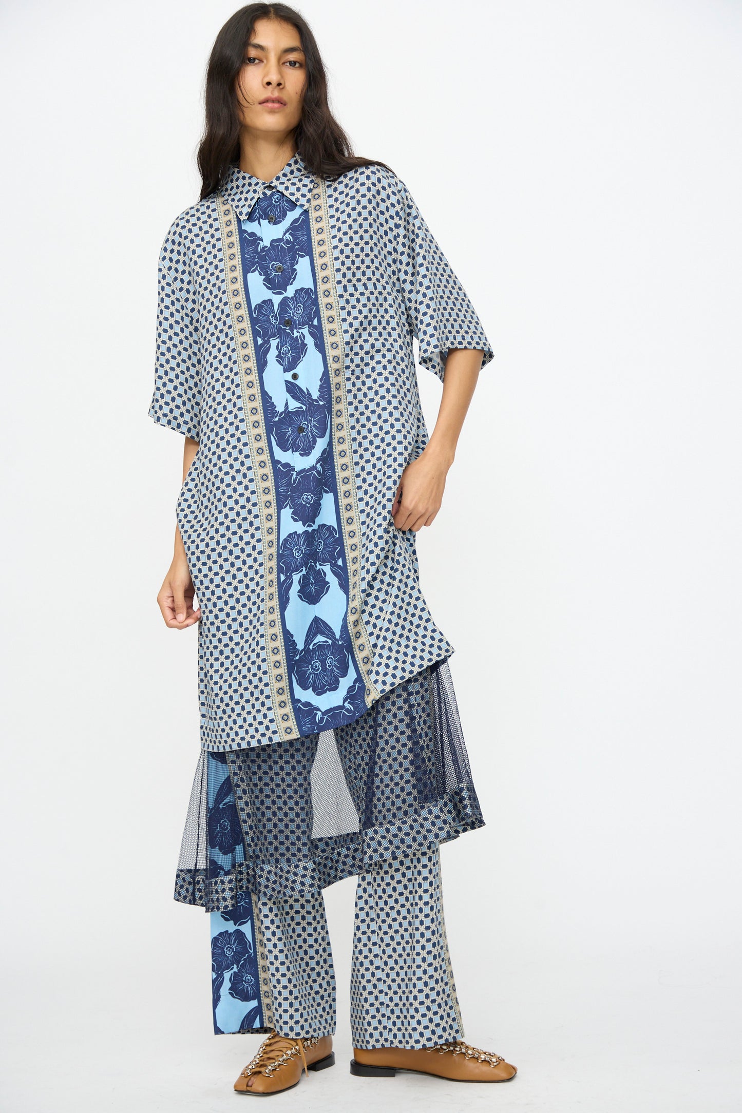 A person wearing the TOGA ARCHIVES Inner Print Dress in Light Blue, featuring floral and geometric prints, over matching wide-leg pants and tan shoes, stands against a plain white background.