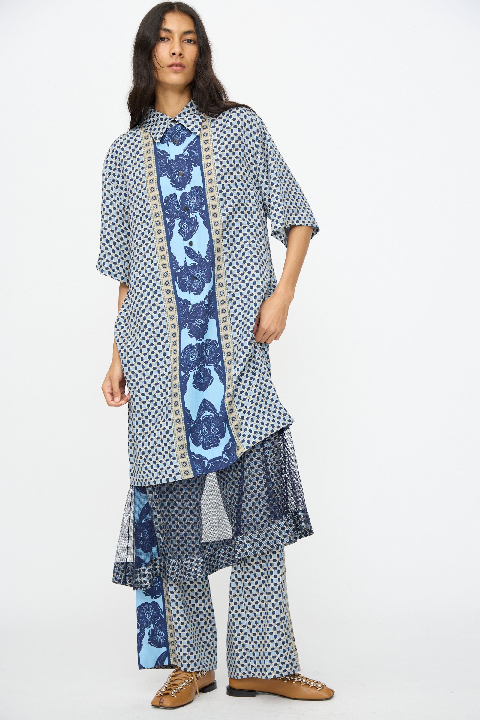A person wearing the TOGA ARCHIVES Inner Print Dress in Light Blue, featuring floral and geometric prints, over matching wide-leg pants and tan shoes, stands against a plain white background.