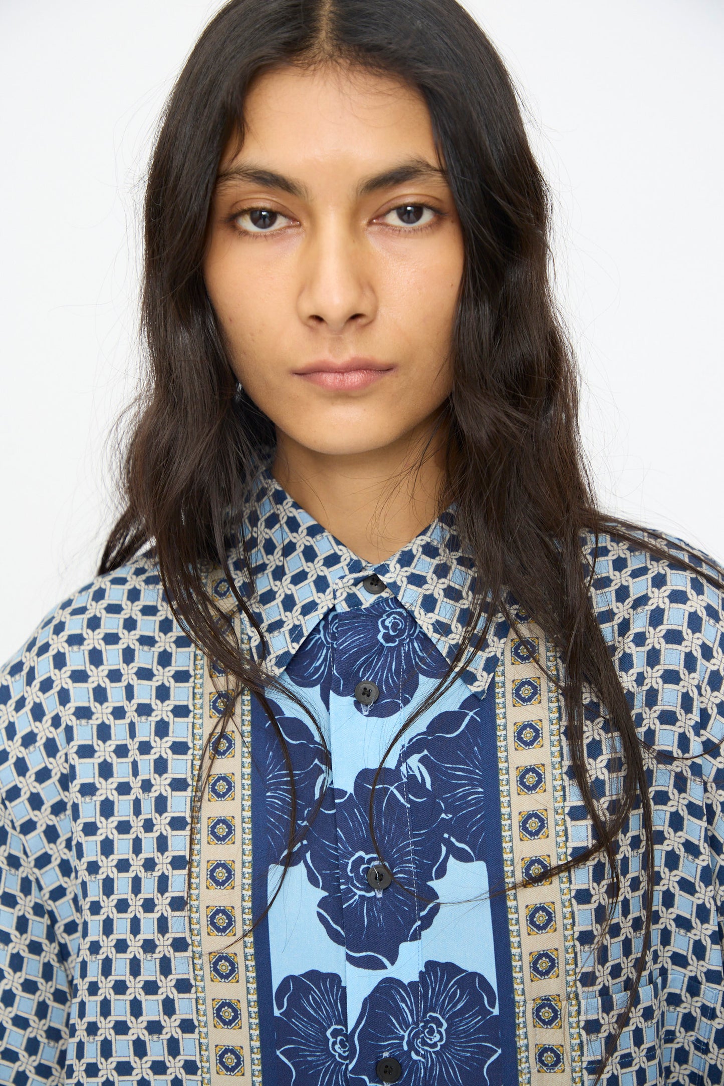 A person with long dark hair models the TOGA ARCHIVES Inner Print Dress in Light Blue, featuring blue geometric and floral patterns, looking directly at the camera against a plain background.