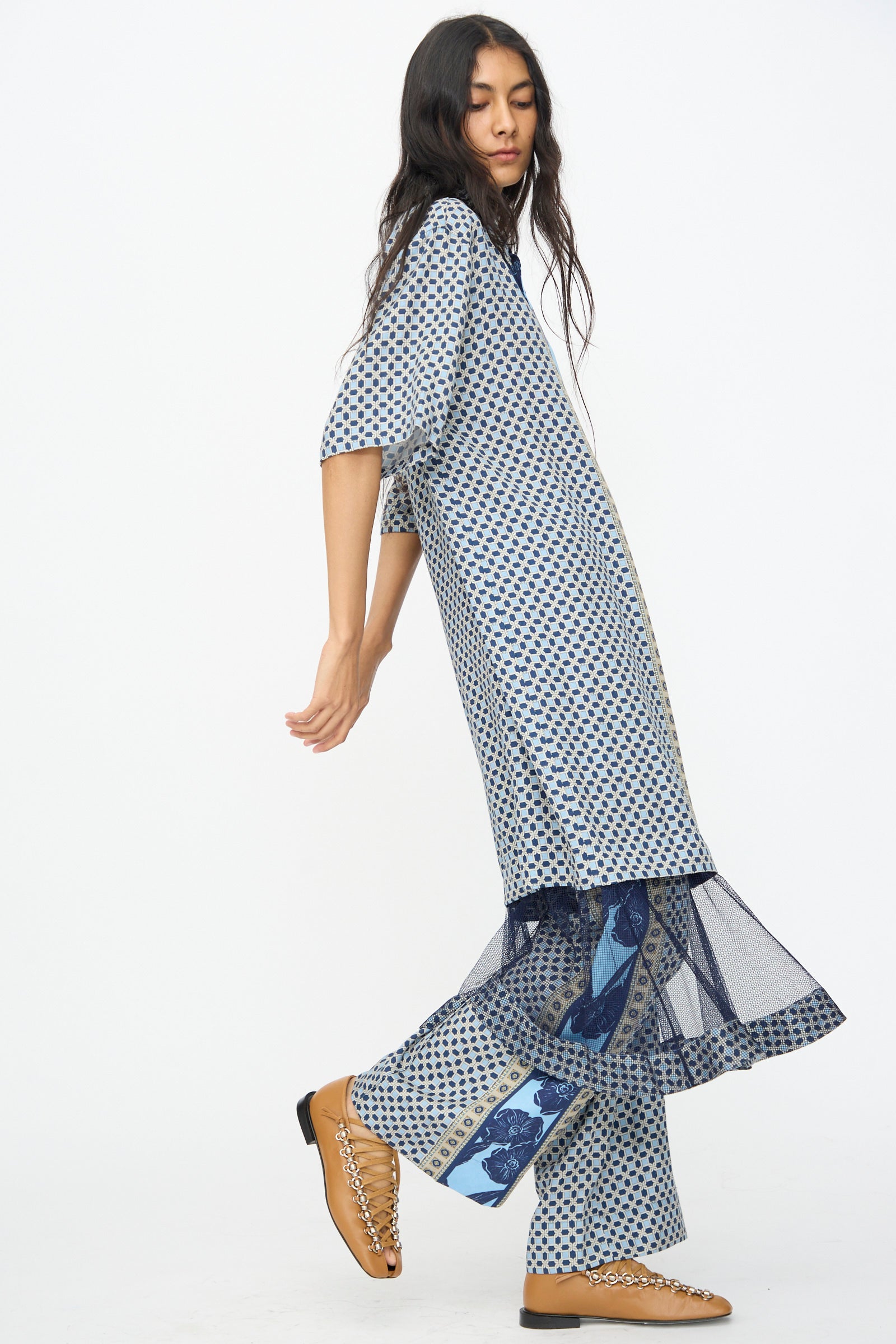 A woman with long hair models the TOGA ARCHIVES Inner Print Dress in Light Blue, a loose-fitting, patterned midi dress with layered ruffles, paired with tan chain-embellished shoes against a plain white background.