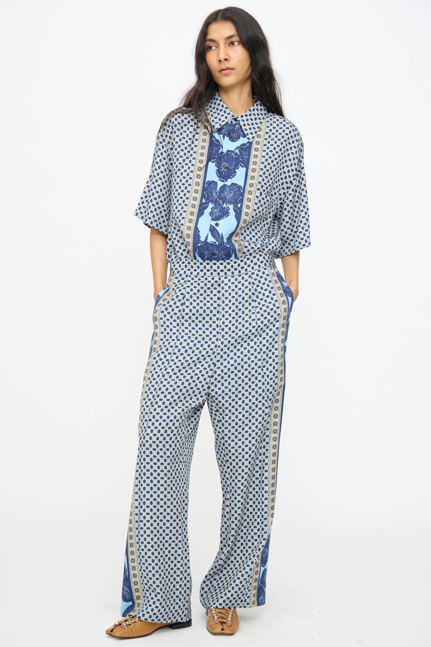 A person wearing TOGA ARCHIVES Inner Print Pant in Light Blue, styled with a loose blue geometric shirt and brown shoes, stands against a plain white background.