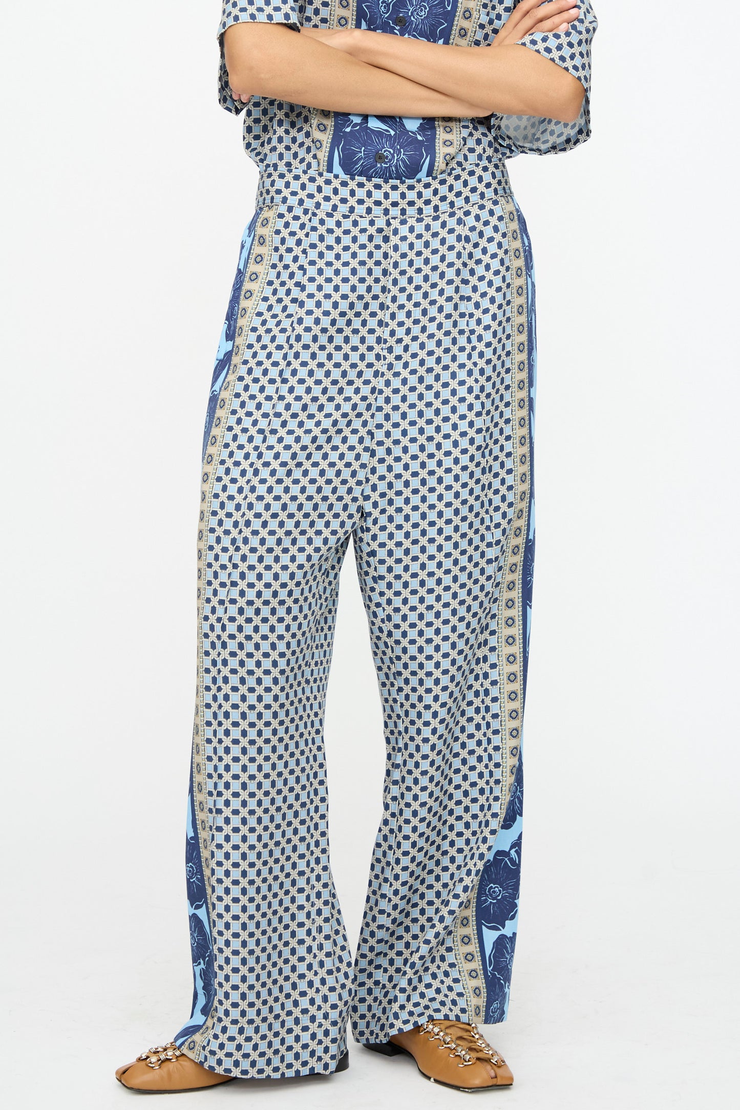 A person stands with arms crossed, wearing TOGA ARCHIVES Inner Print Pant in Light Blue and tan chain loafers, cropped to show only the lower body against a plain background.