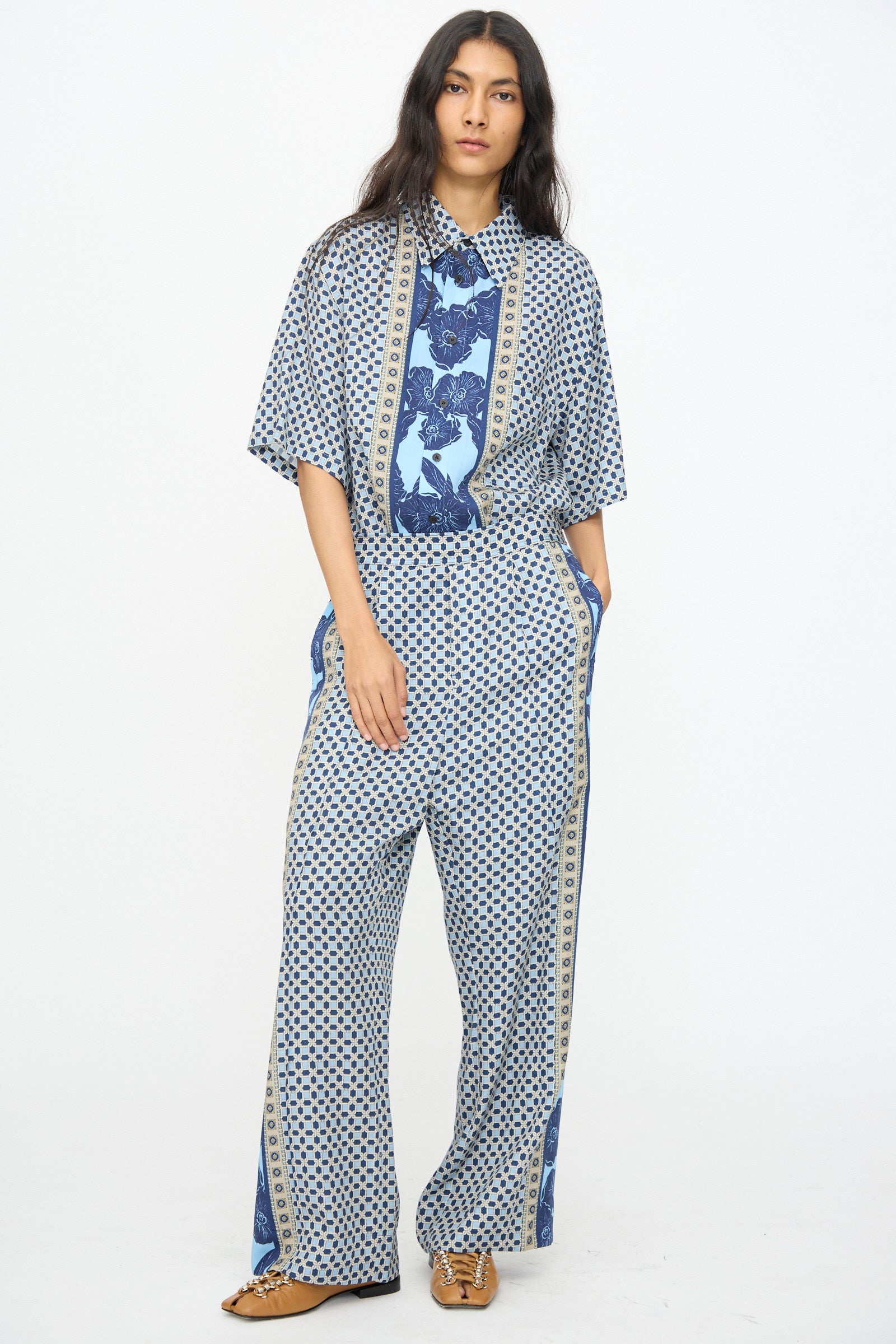A person stands against a plain background wearing TOGA ARCHIVES’ Inner Print Pant in Light Blue with a loose-fitting geometric shirt and tan shoes—an effortlessly chic Japanese-made look.