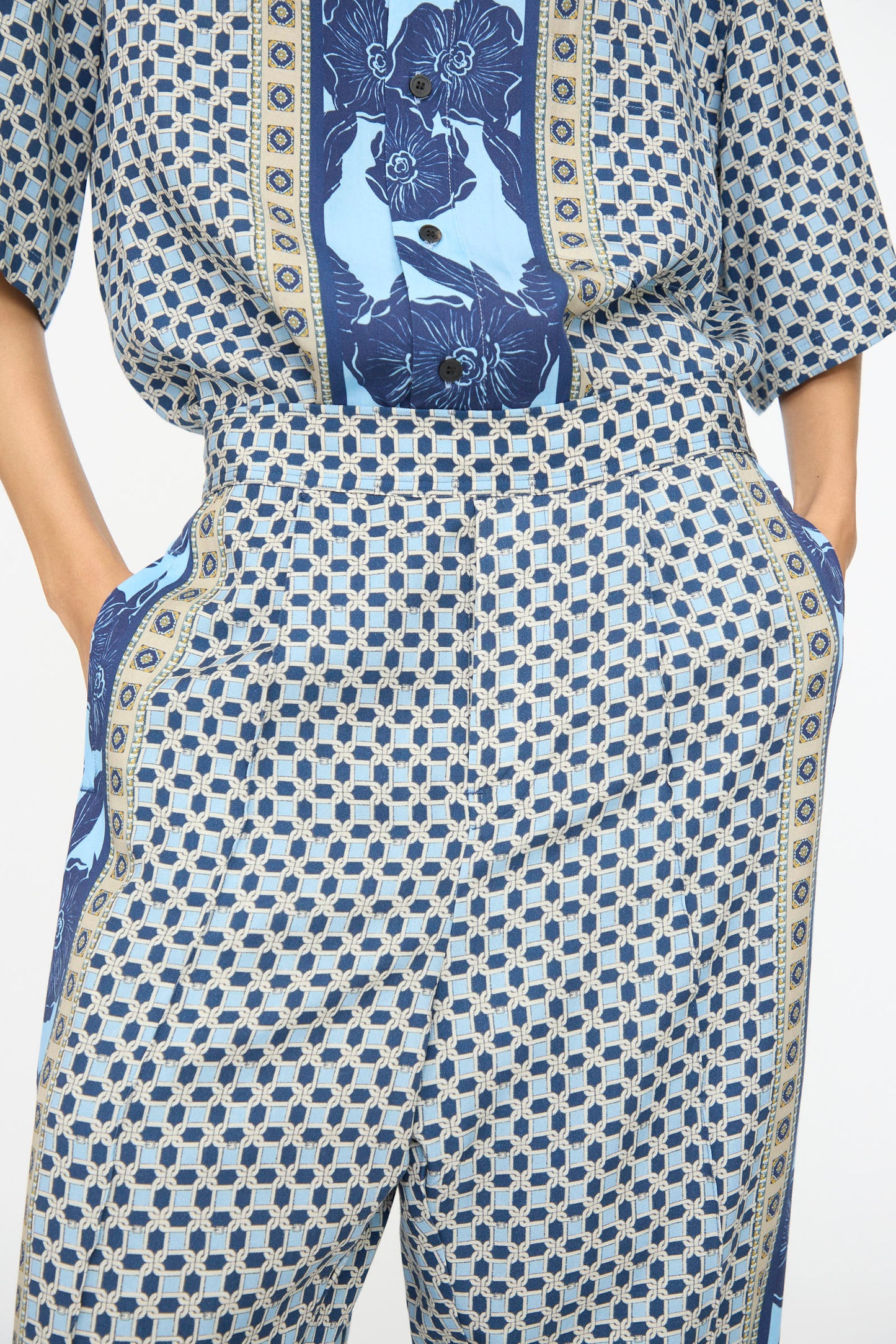 Model wearing TOGA ARCHIVES Inner Print Pant in Light Blue with hands in pockets, styled with a matching shirt, both showcasing blue geometric and floral patterns.