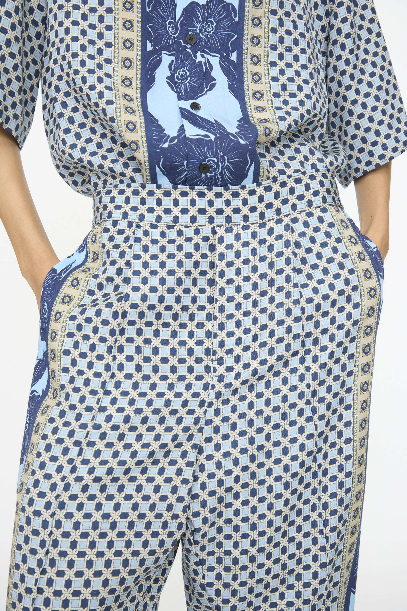 Model wearing TOGA ARCHIVES Inner Print Pant in Light Blue with hands in pockets, styled with a matching shirt, both showcasing blue geometric and floral patterns.